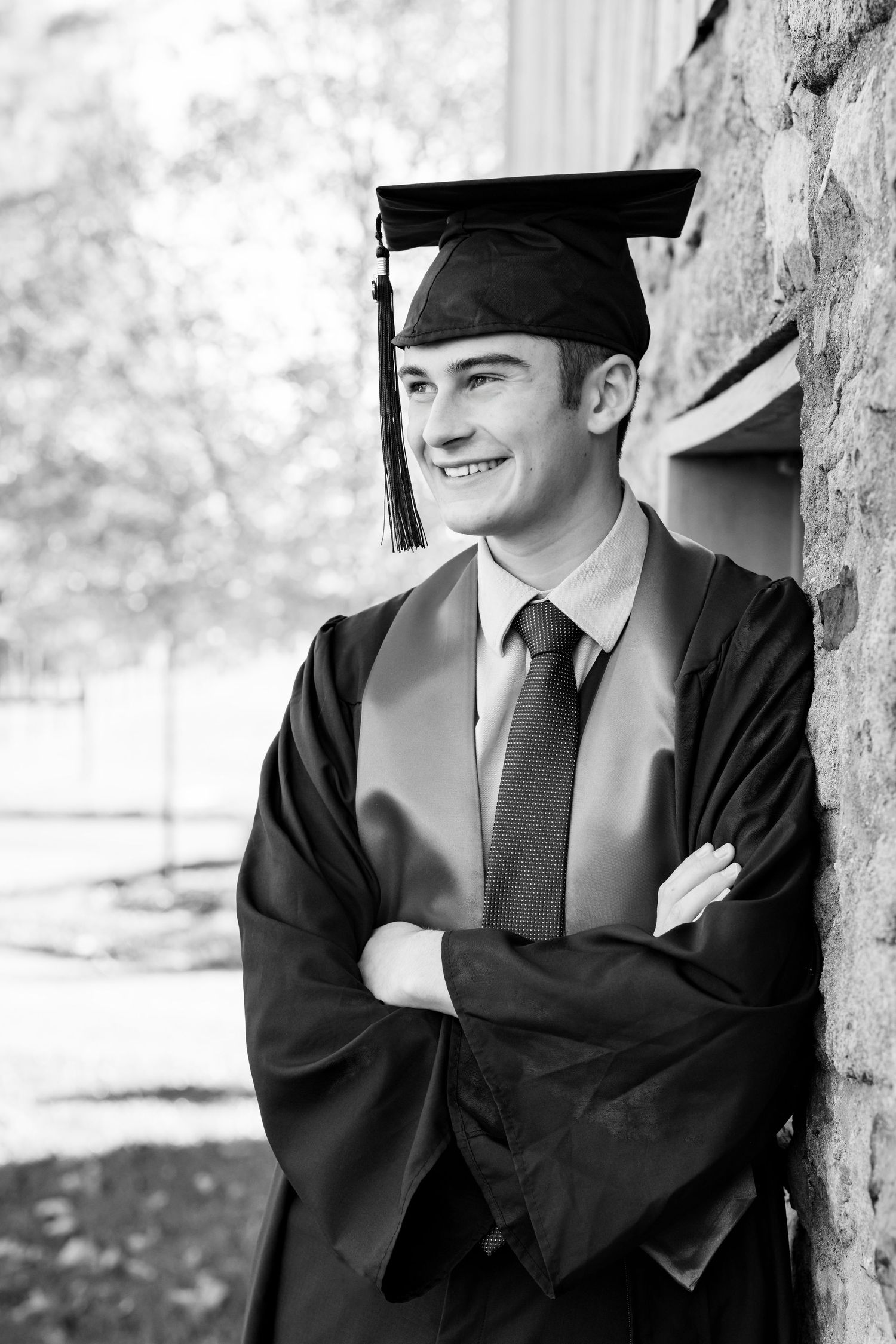 A graduate in cap and gown poses outdoors against a stone wall in black and white photography.