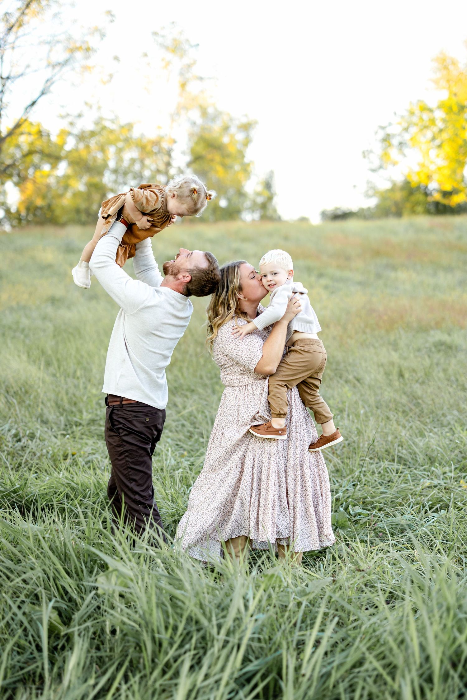 Happy family with two babies kissing in a green meadow during a warm autumn day outdoor photo session.