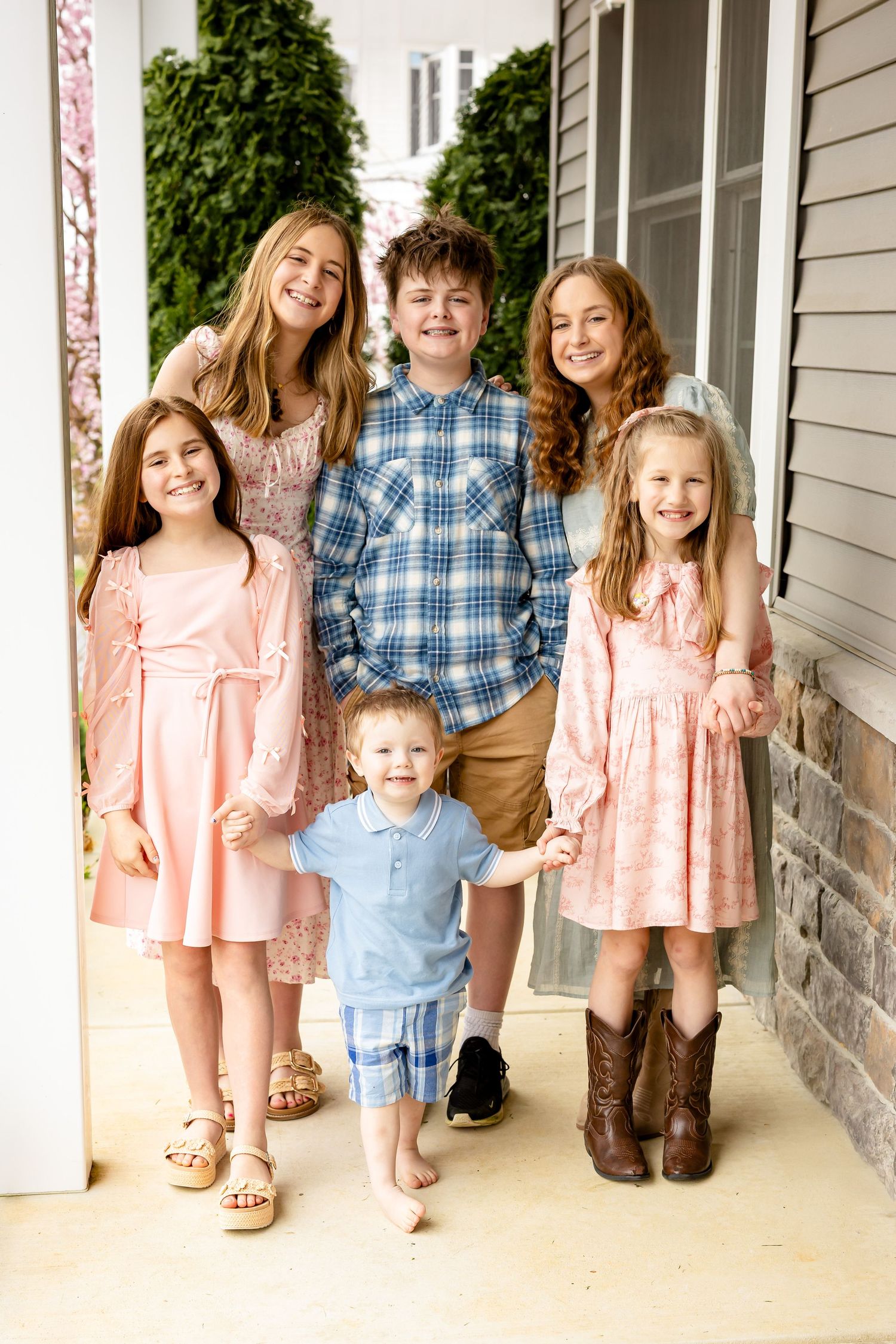 Six children in pastel outfits pose together smiling on a home porch for a cheerful family portrait.