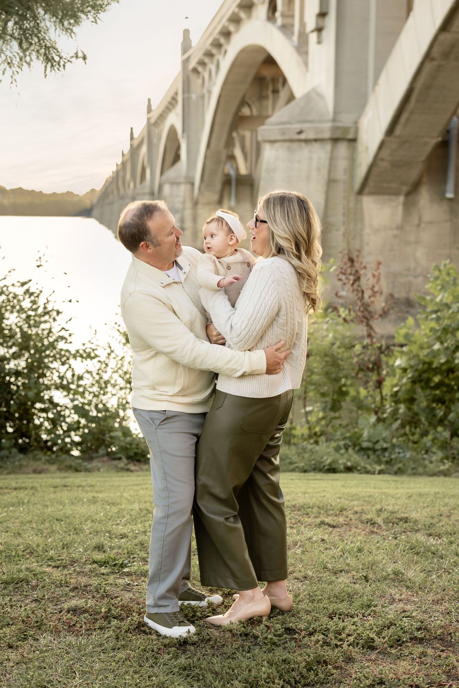 A couple holds their baby by a river bridge, wearing cream sweaters and olive pants in a warm outdoor setting.