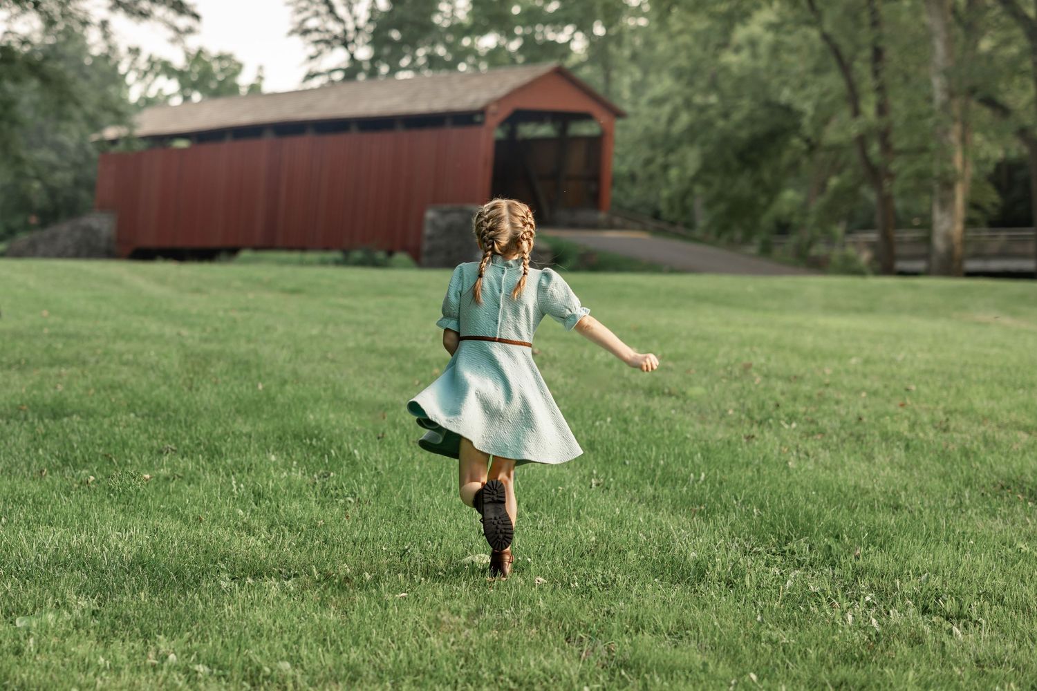A young girl in a teal dress and boots runs across a green lawn toward a red covered bridge.