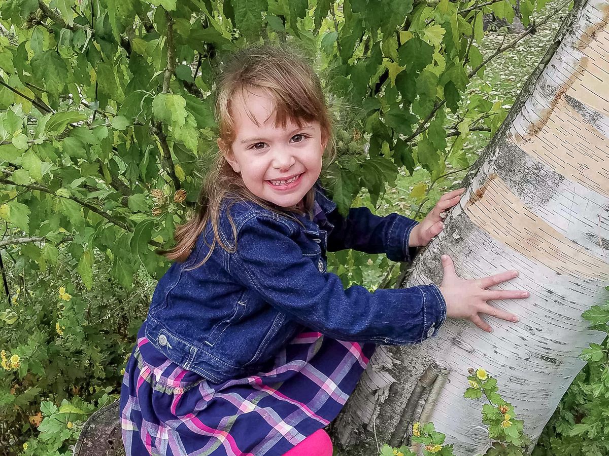A cute young girl poses playfully next to a birch tree trunk in a leafy outdoor setting while wearing a navy blue and plaid outfit.