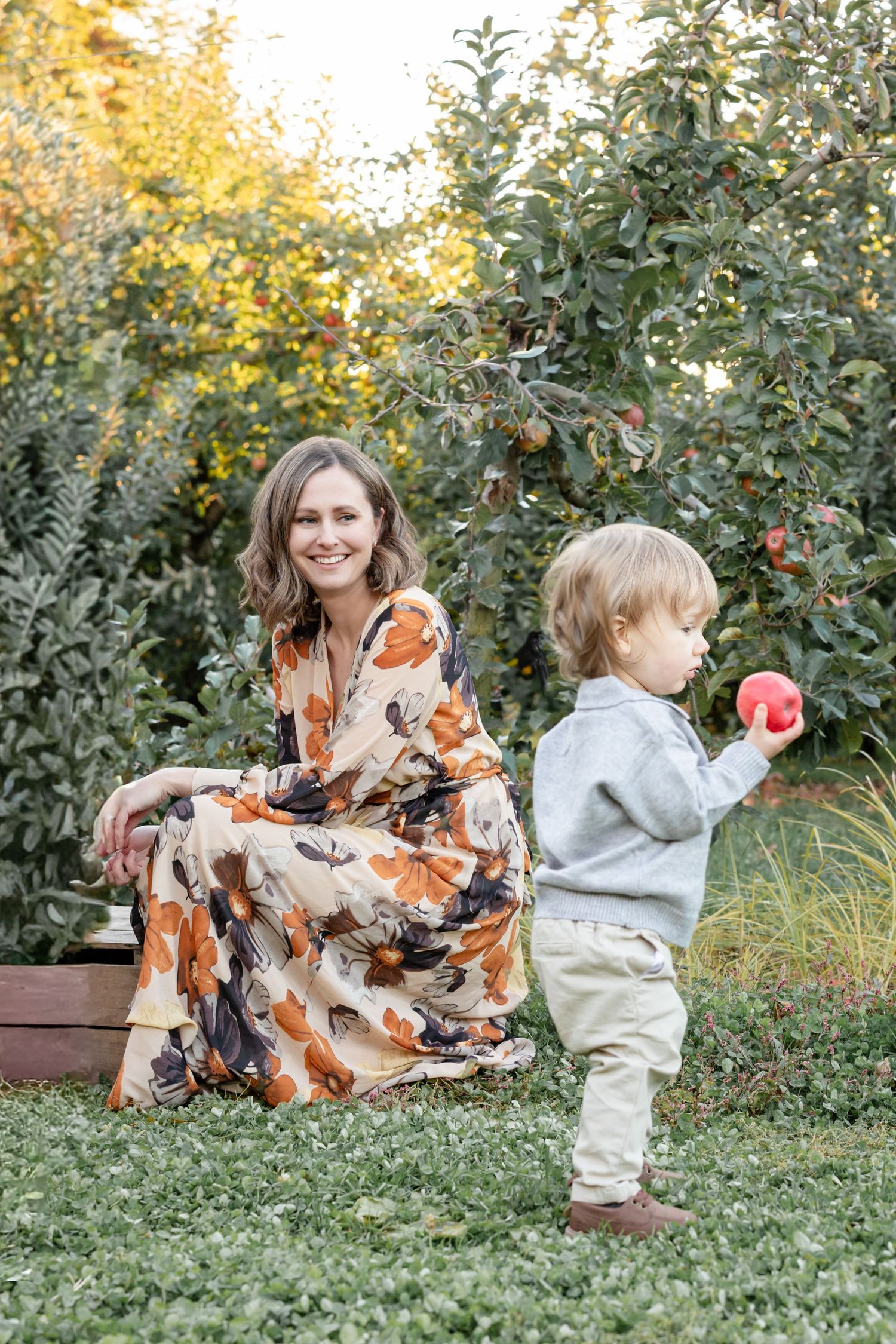 A mother in a refined floral maxi dress enjoys the orchard with her young son dressed in a grey sweater and beige pants