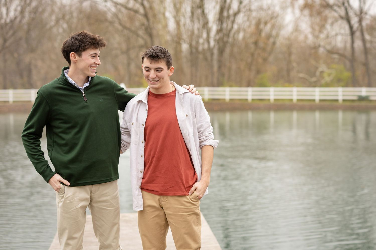 Two senior boys in casual attire walk together and laugh by a serene lake on an autumn afternoon.