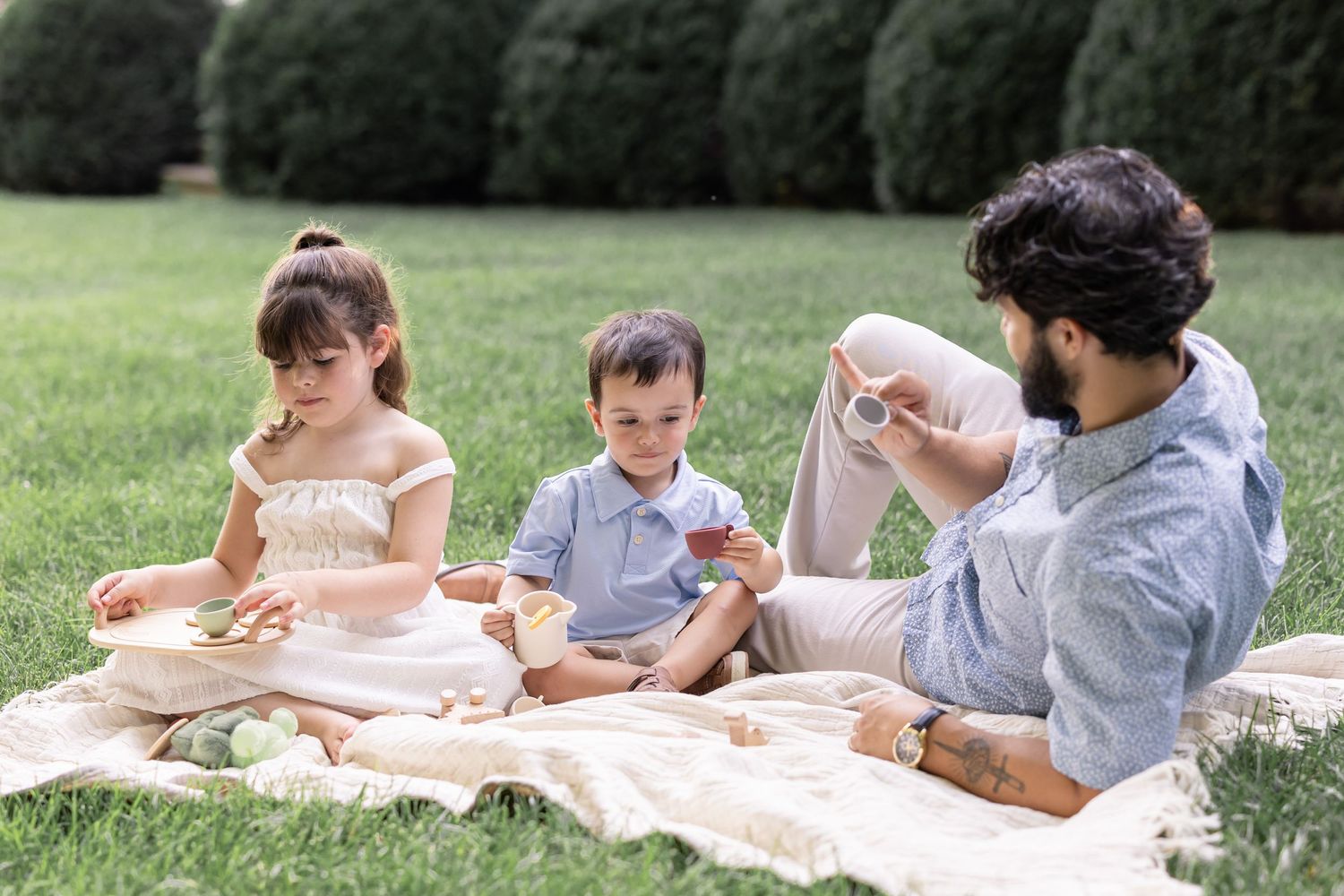 In a candid family photo, a father enjoys a quaint tea party with his two young children on a lush green lawn in Lancaster, PA.