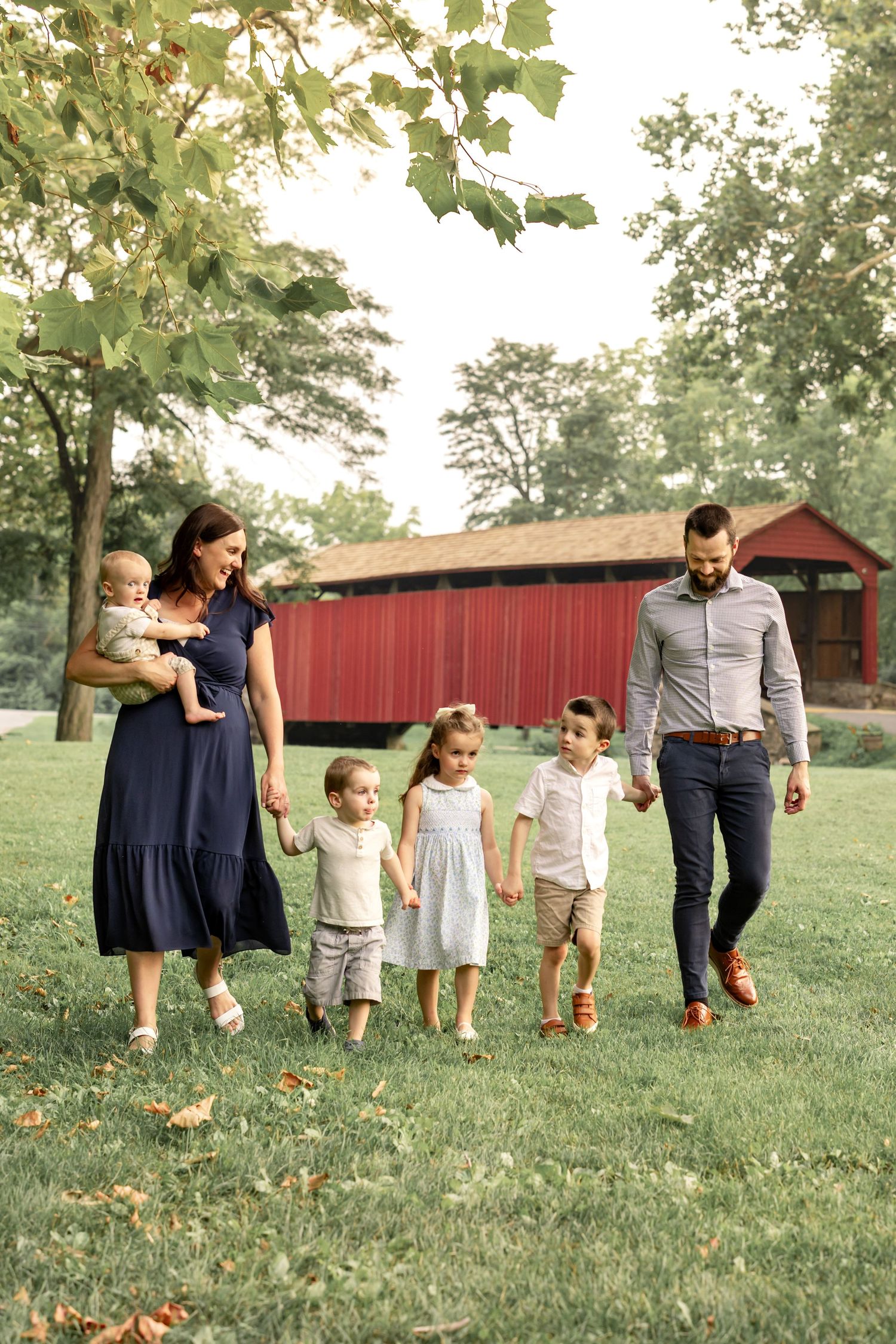 Family of six walking on green grass near a red covered bridge, surrounded by lush trees on a sunny day.