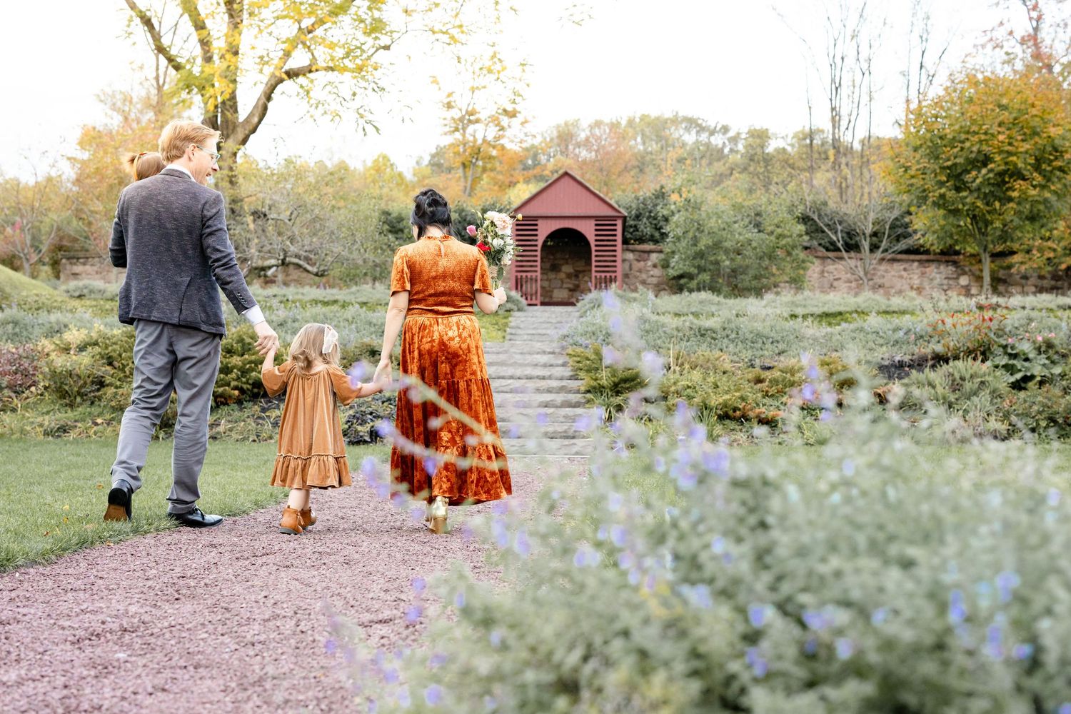 Family of three walking through a lush garden path toward a red brick gazebo in autumn.