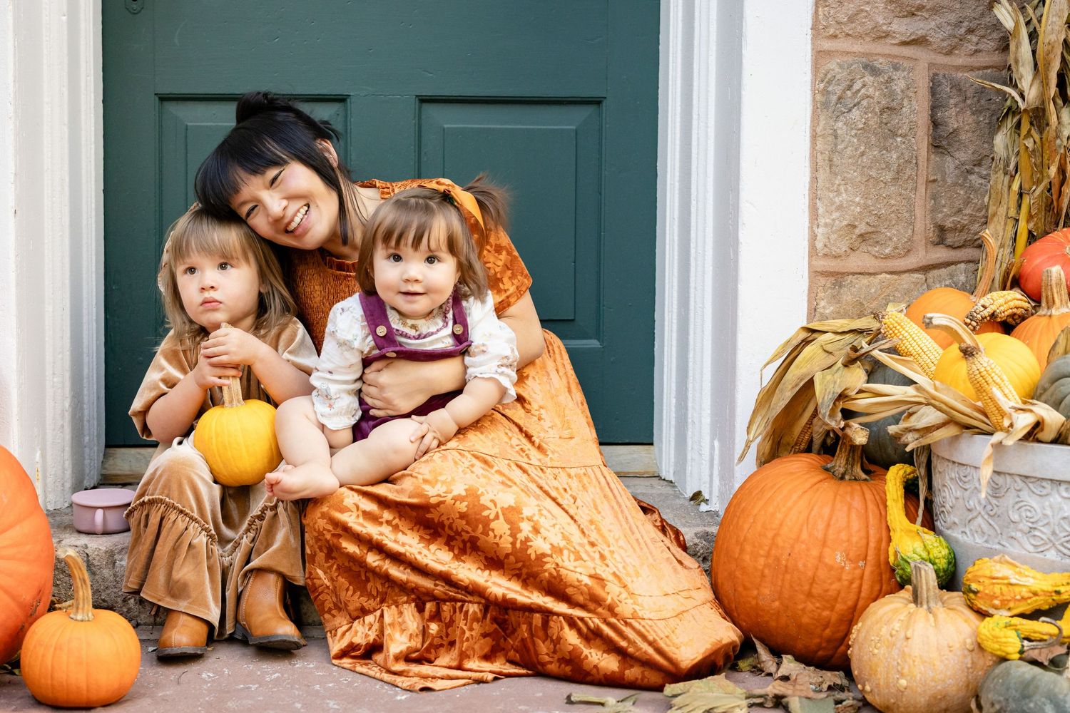 Woman and two children sitting on porch steps surrounded by pumpkins and fall decorations for Halloween.