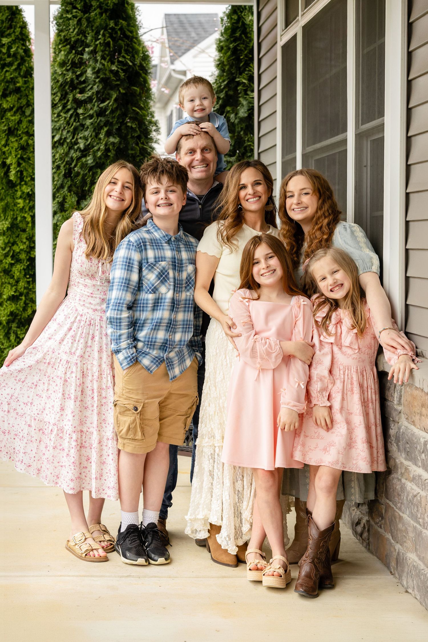 Large family of eight posing together on a home porch, dressed in coordinating pastel outfits, smiling warmly.