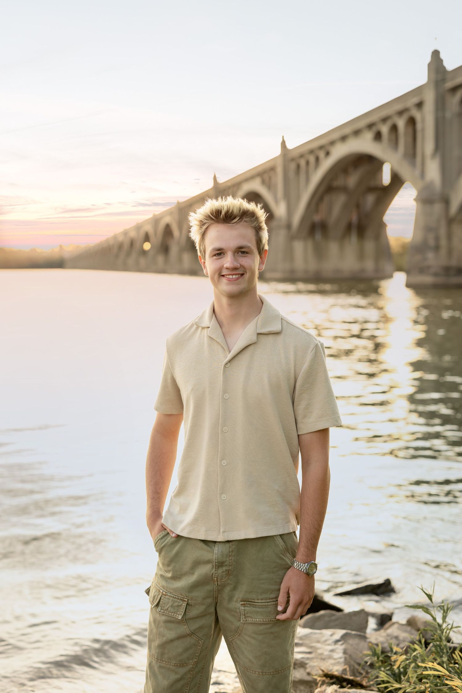 Outdoor portrait of a young man at sunset by a stone arch bridge over water with golden light reflecting on the rippling surface.