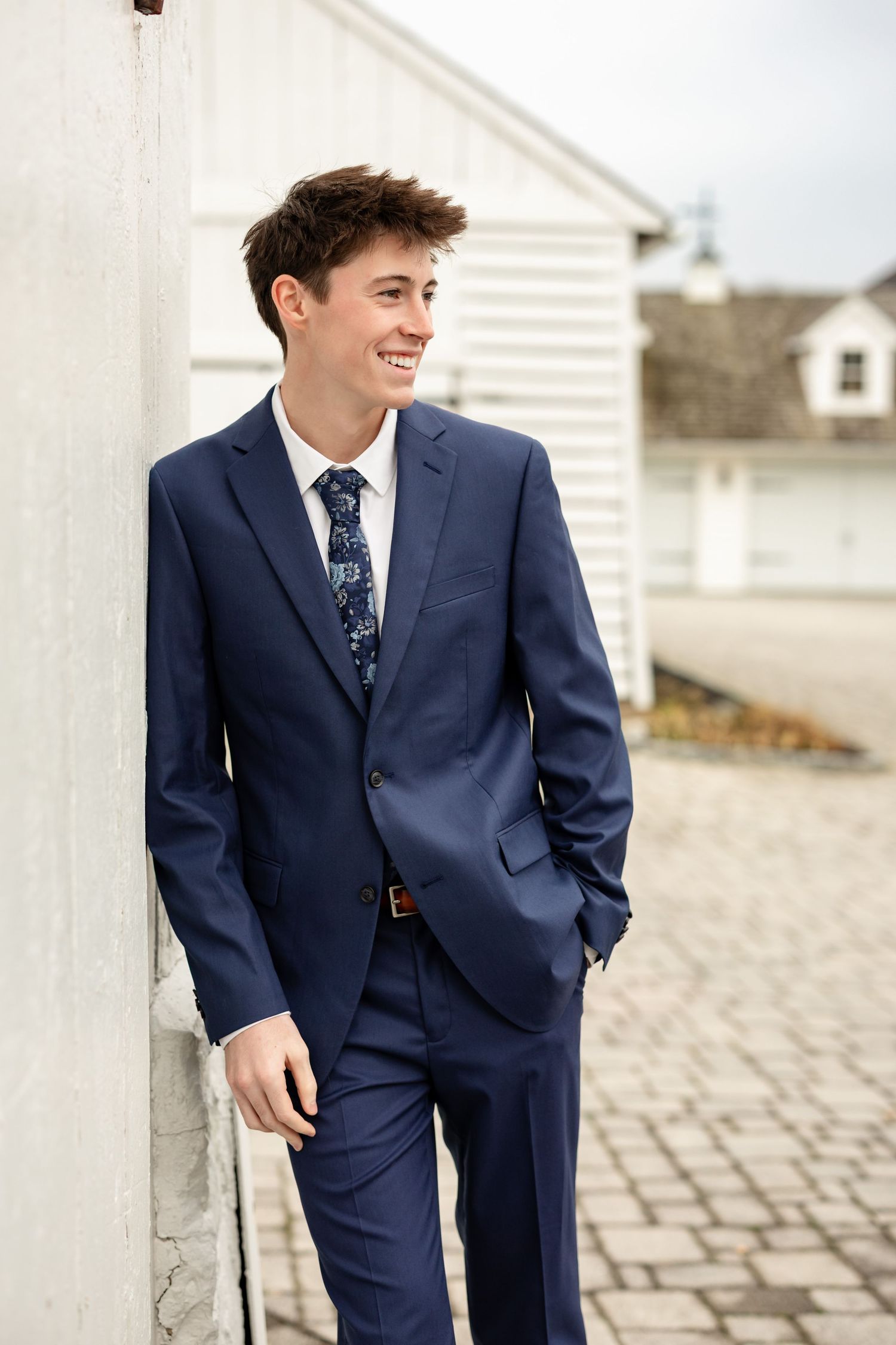 A high school senior guy in a navy suit and patterned tie leans against a white pillar while smiling outdoors.