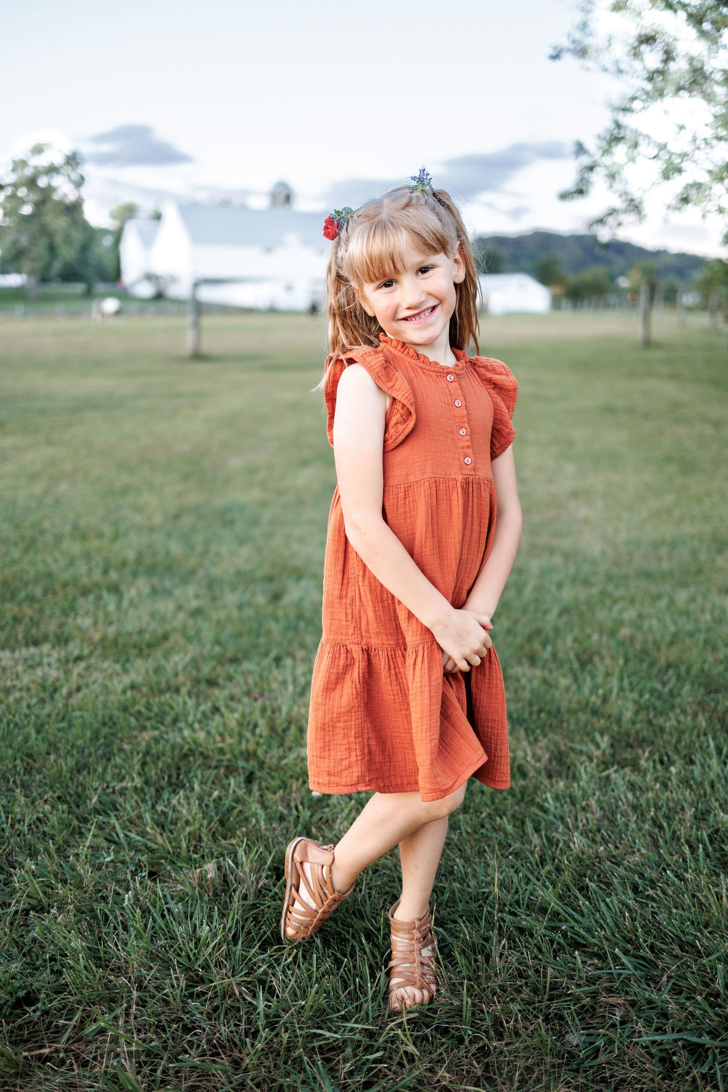 A smiling young girl in an orange ruffle dress and sandals stands on green grass near a white barn.