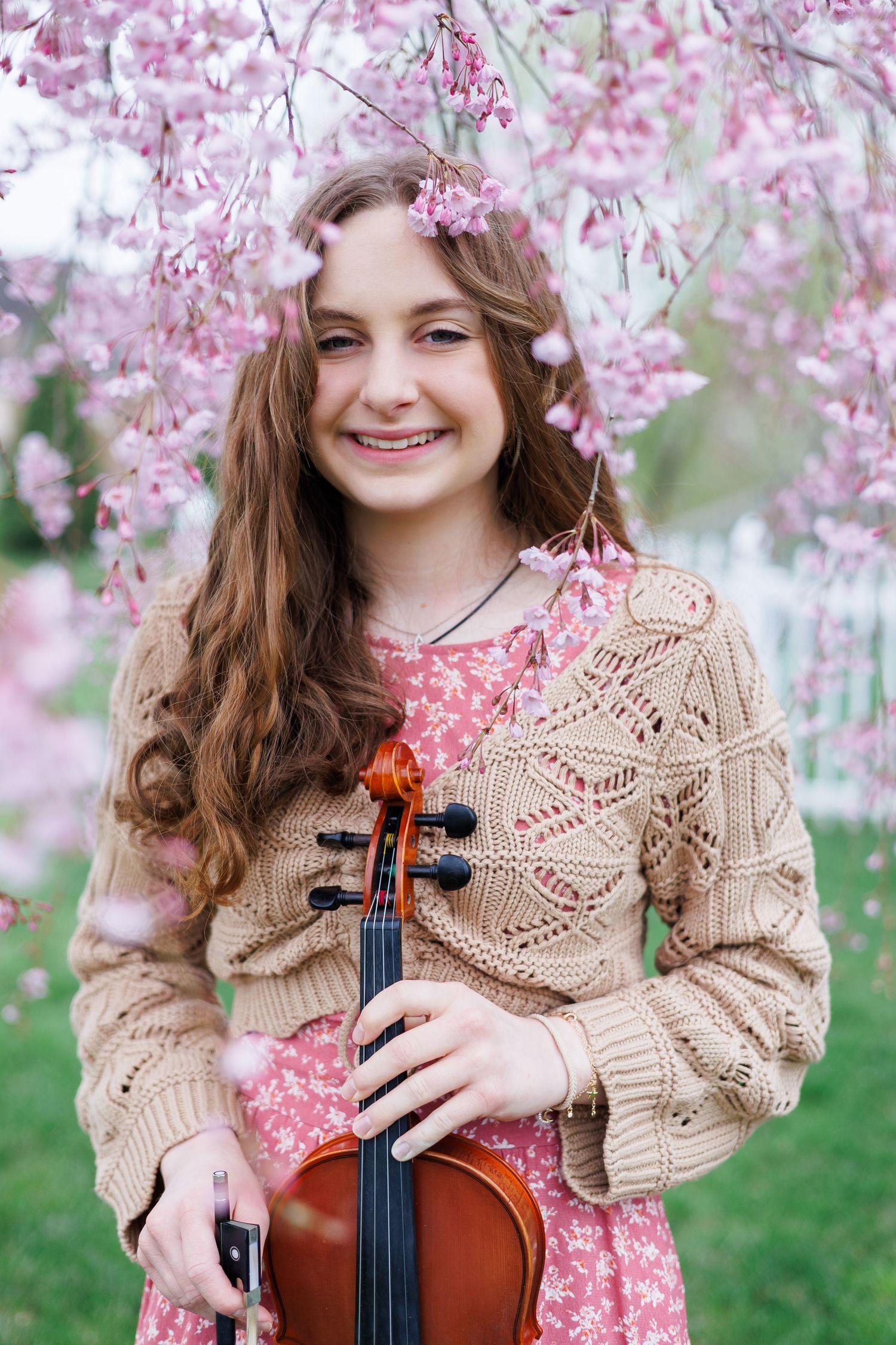 A high school senior girl in a beige knit sweater poses with a violin under blooming pink cherry blossoms in spring.