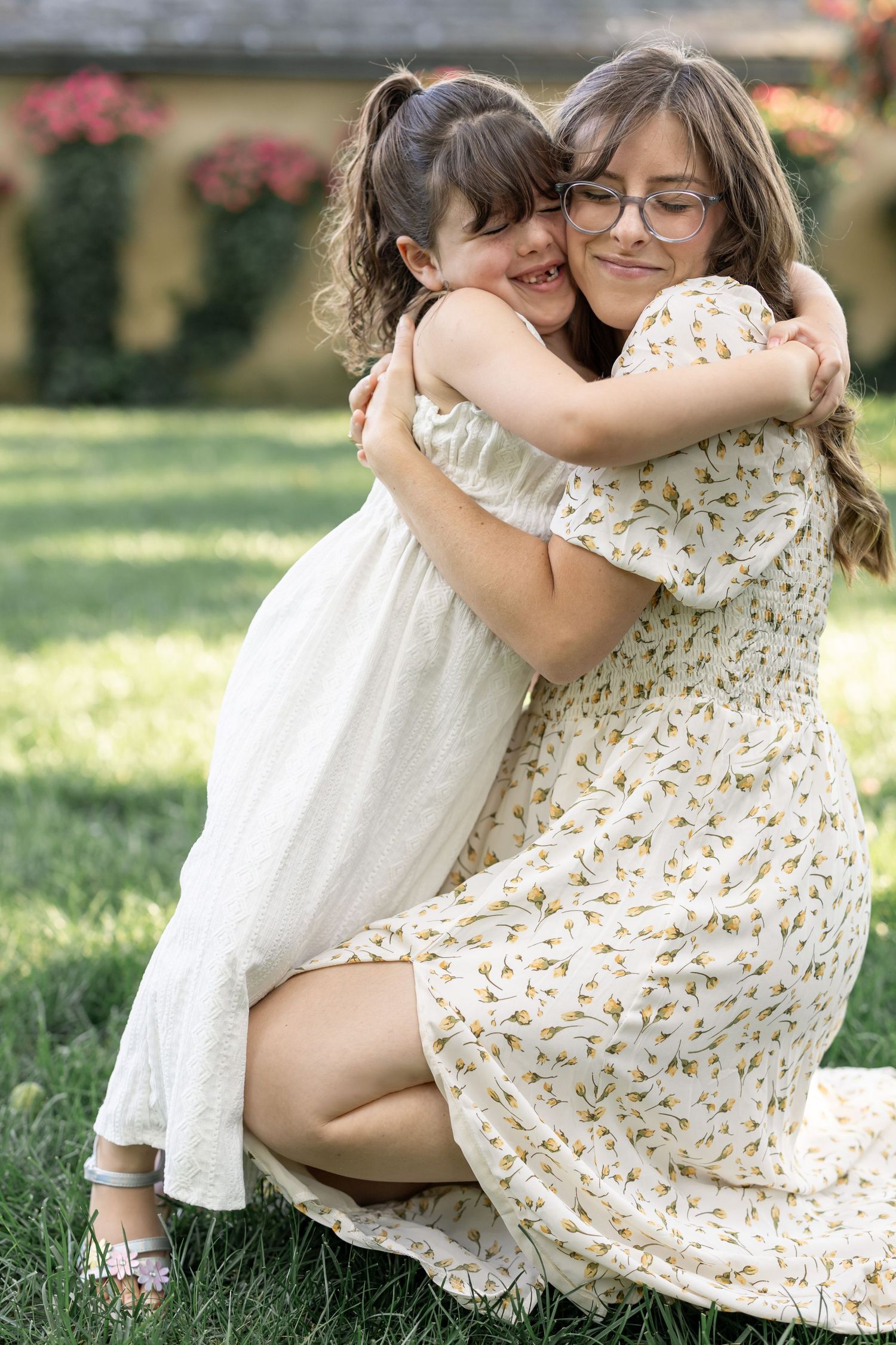 Two girls in floral dresses share a warm hug outdoors on green grass with blooming flowers in the background.