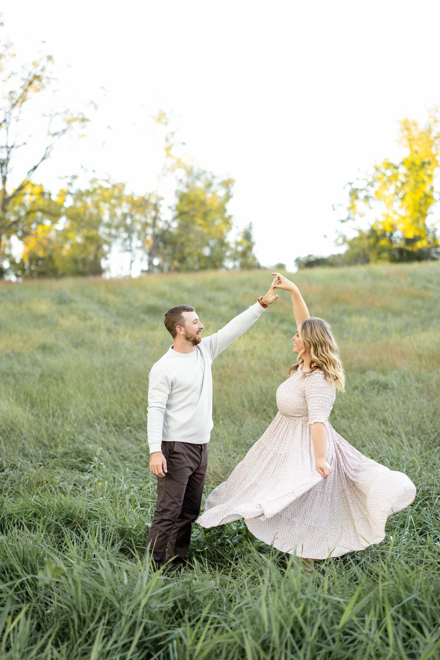 A couple dances in a green meadow, man spinning woman in a flowing blush dress at golden hour.