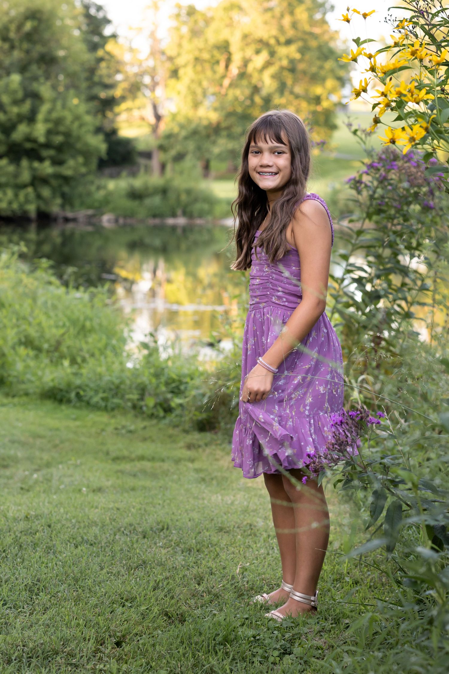 A little girl in a purple dress plays outdoors near a pond on a sunny autumn afternoon.
