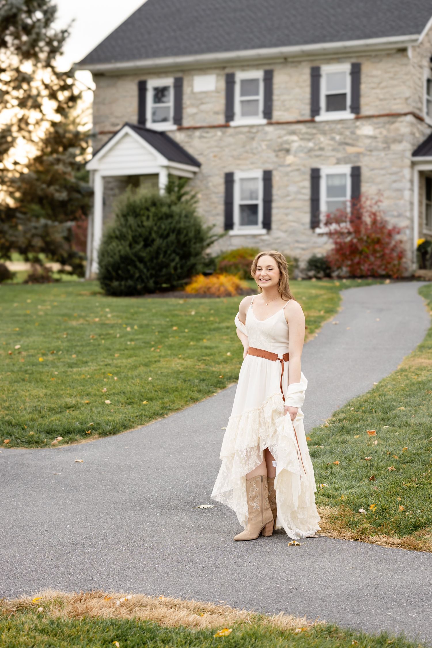 A high school senior girl in an elegant cream-colored dress sits gracefully on a winding driveway with stone houses in the background.