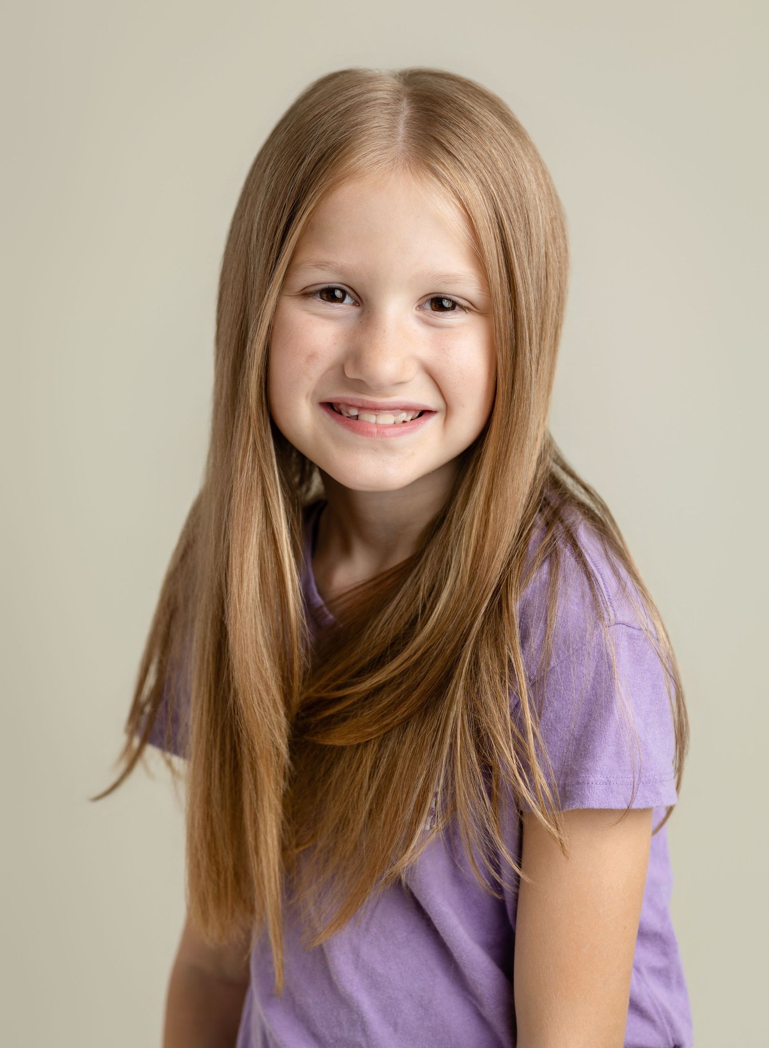 Smiling young girl with long blonde hair wearing a purple t-shirt poses for a portrait photo.