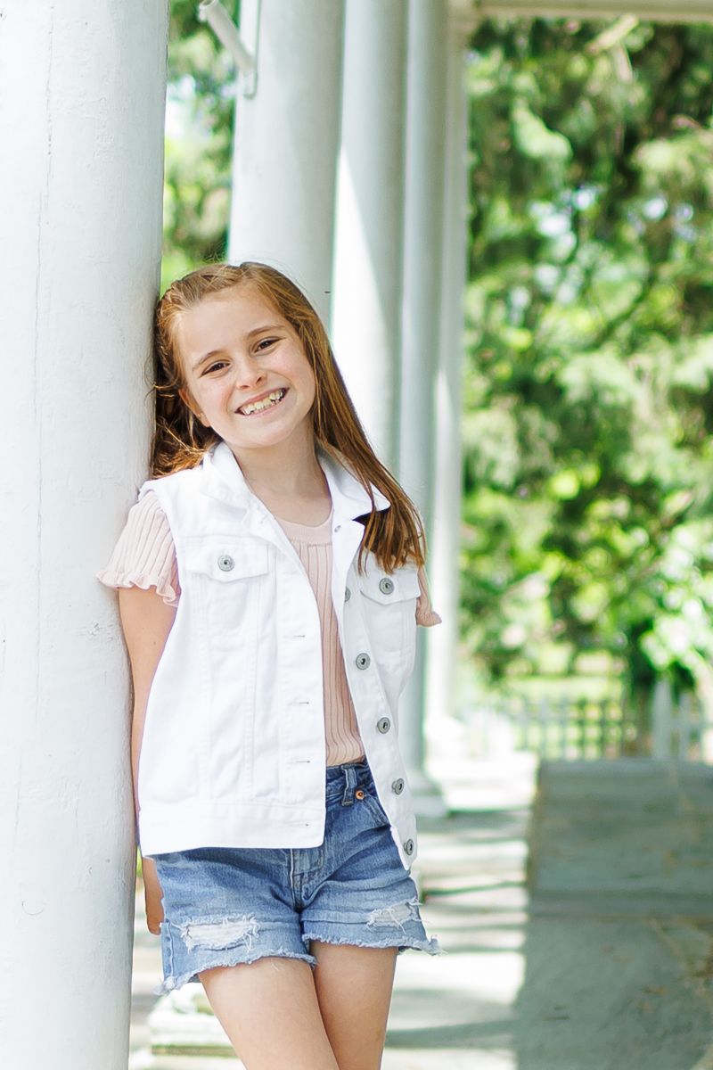 A family milestone session in Gap, PA at White Chimneys Estate. A young girl in shorts and a white vest leans on a column on a grand porch.