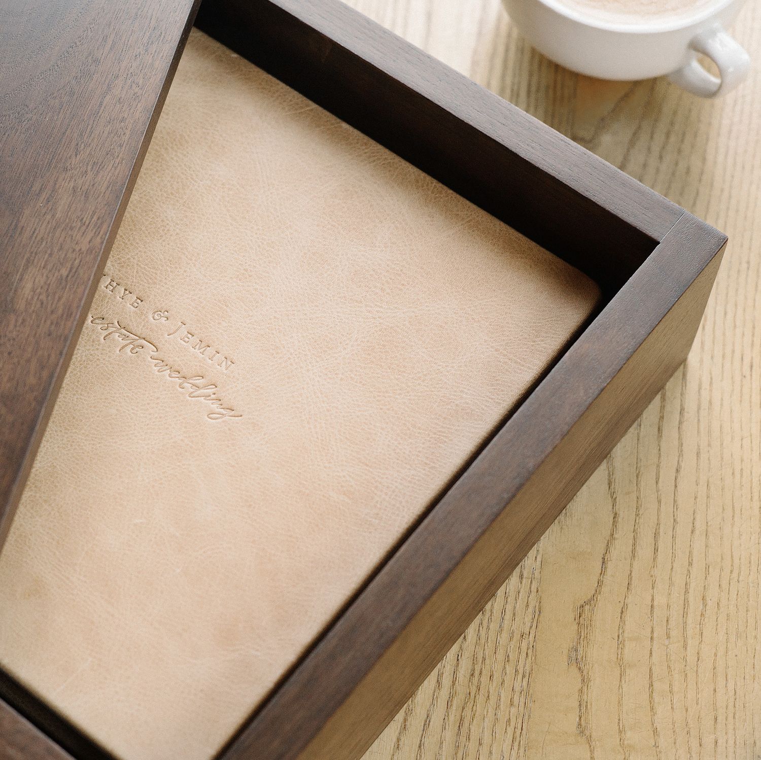 A wooden box with a dark brown frame and beige interior sits on a light wooden surface with a coffee cup nearby.