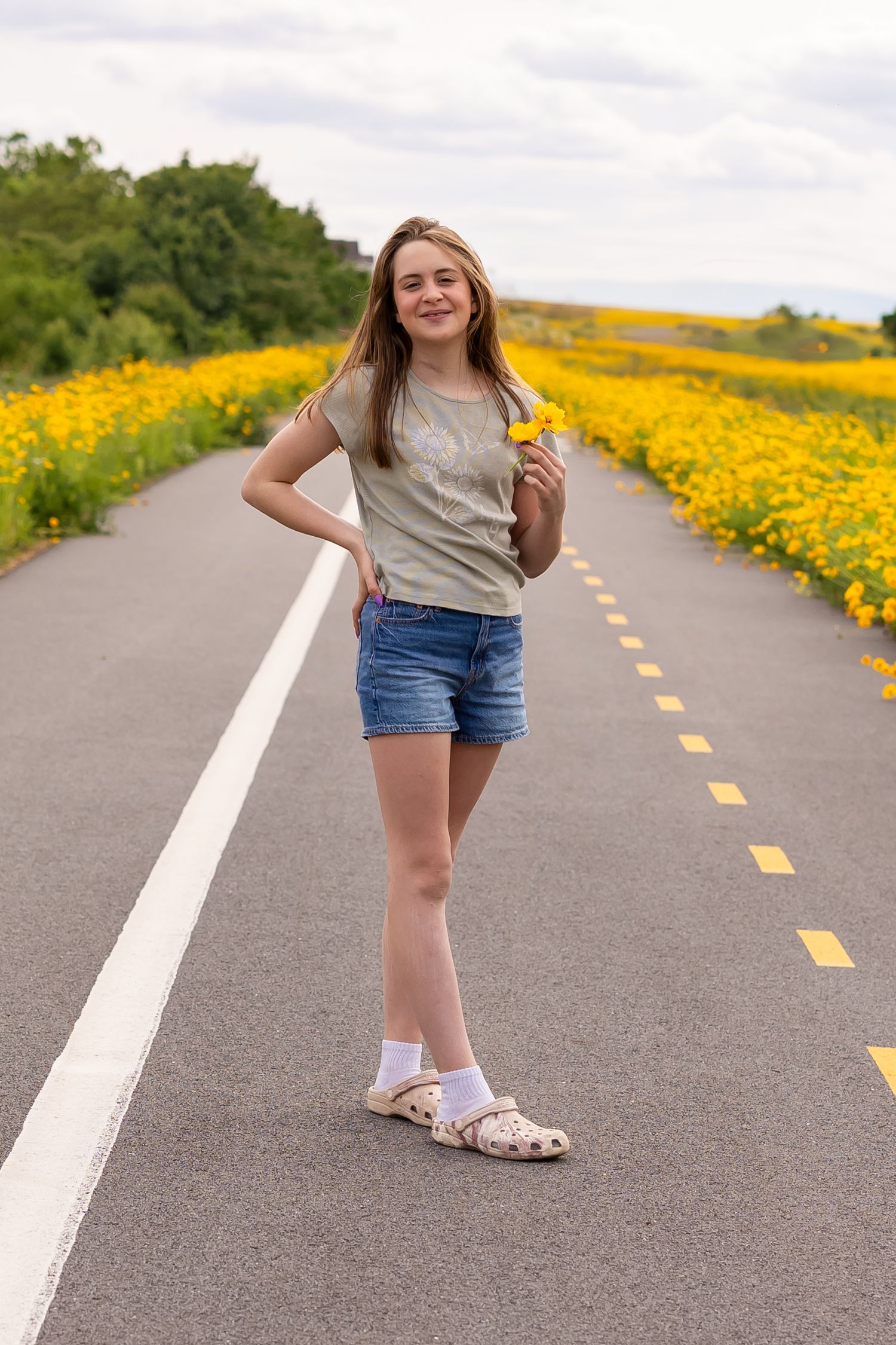 A young lady stands on a paved path bordered by vibrant yellow wildflowers on a summer day.