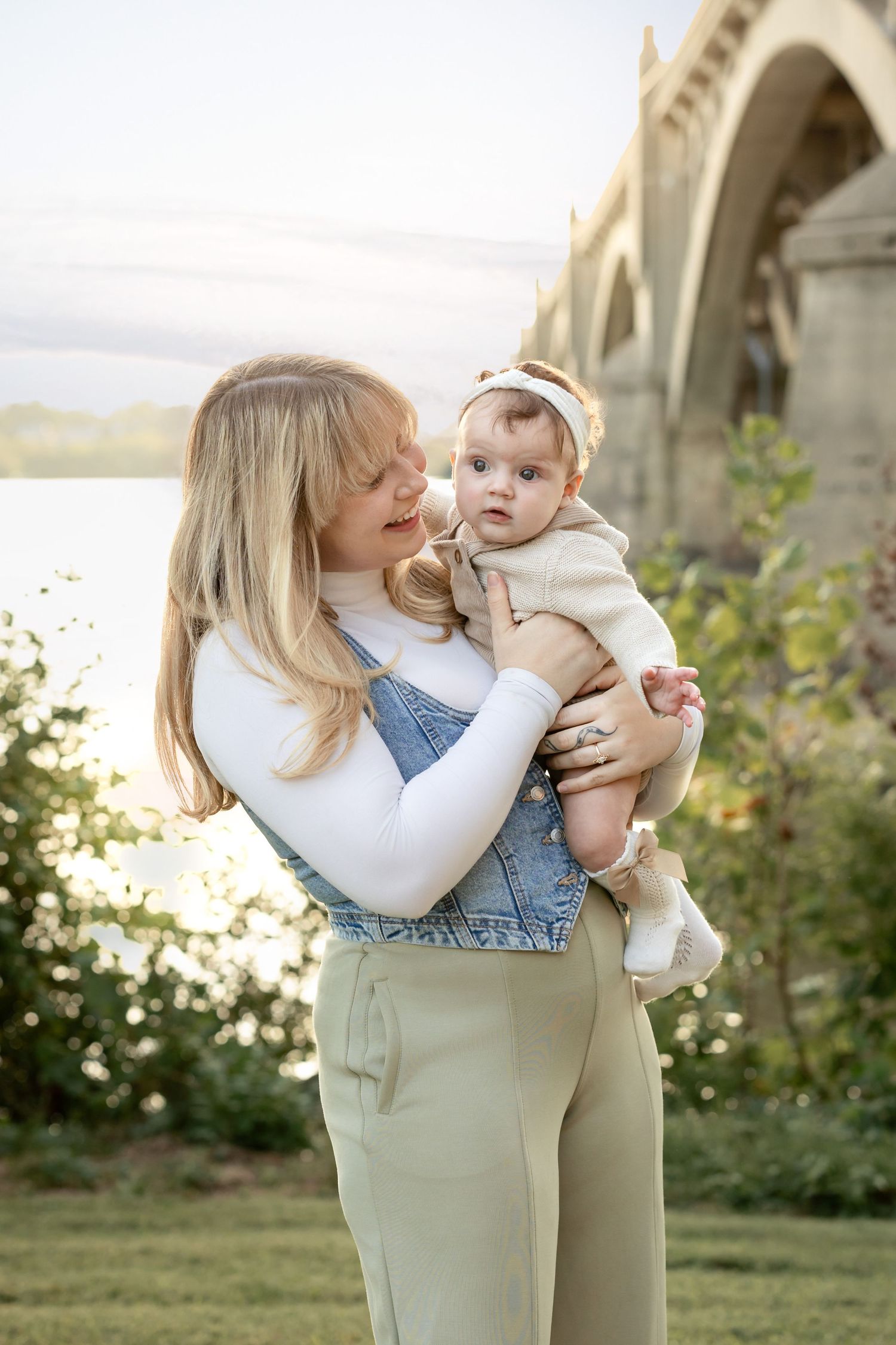 A blonde woman in denim overalls holds a baby girl outdoors near a river with an arched bridge backdrop.
