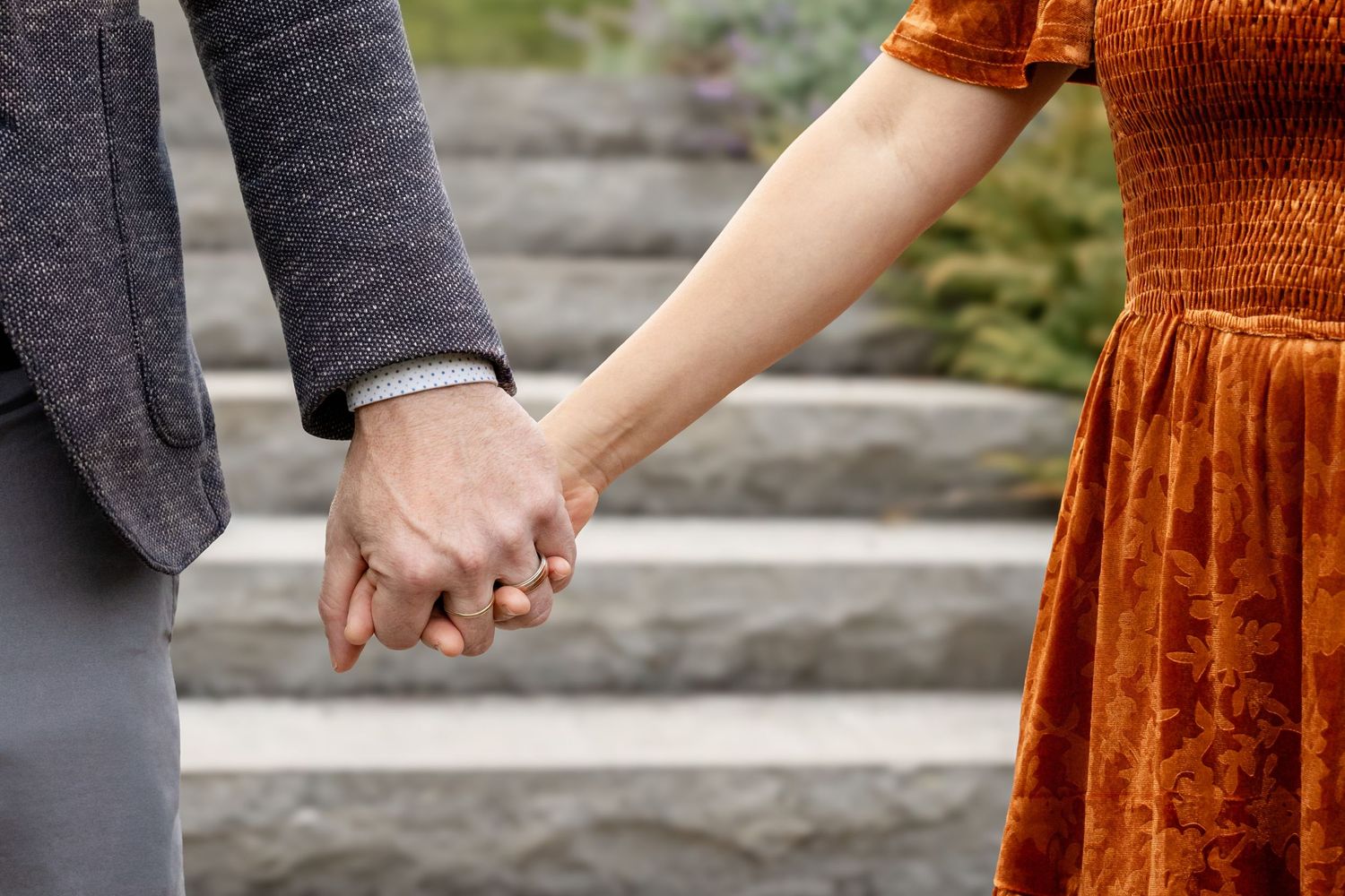 Couple holding hands outdoors, man in gray suit and woman in rust orange velvet dress, standing on stone steps.