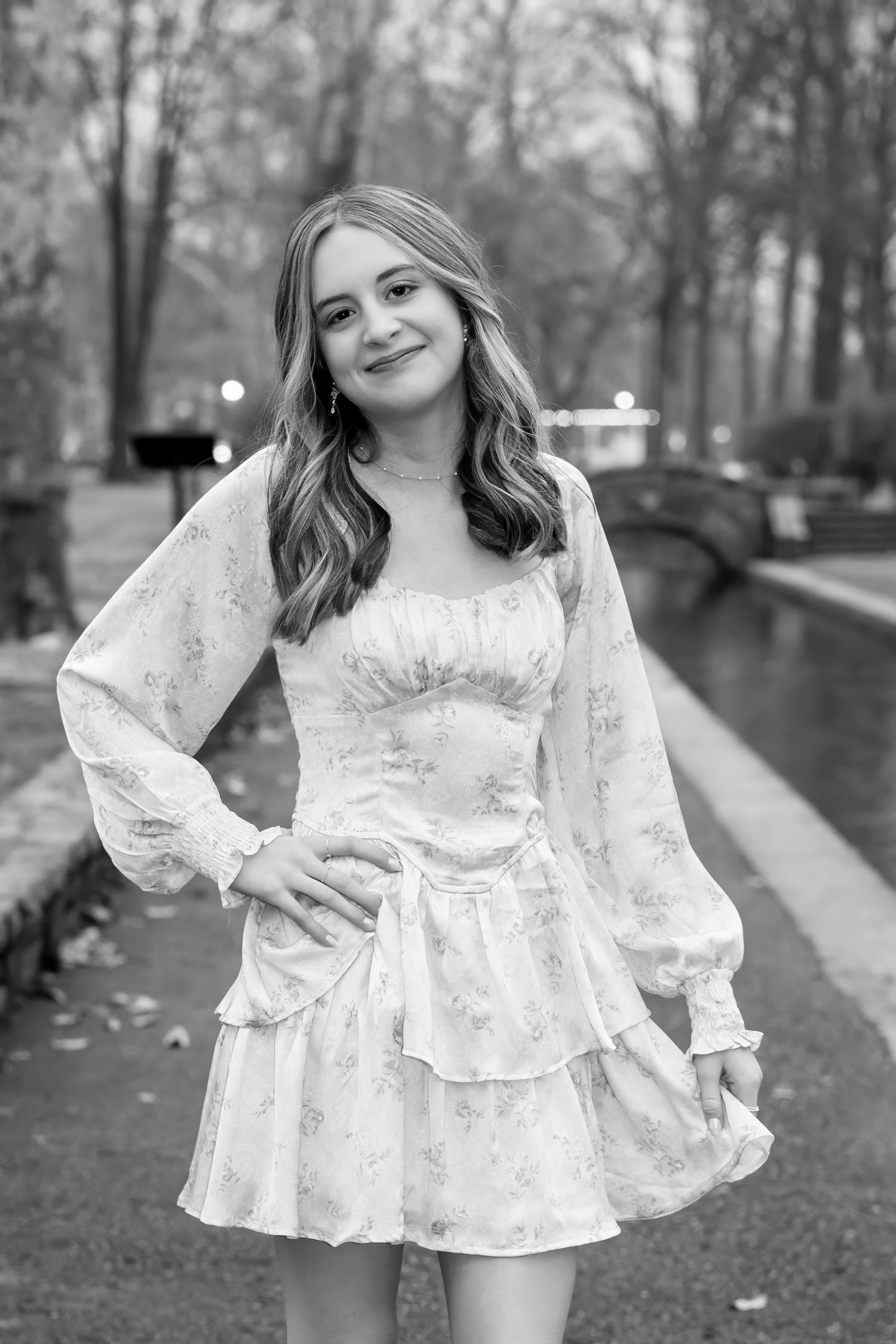 A high school senior in an elegant floral dress poses outdoors in a park setting captured in black and white photography.