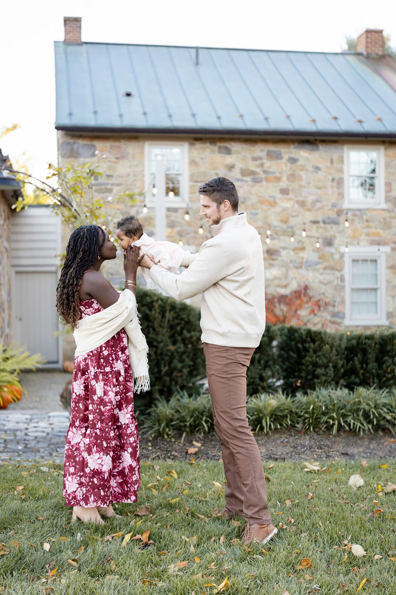 A happy interracial couple holds their baby outdoors in front of a stone house during autumn.