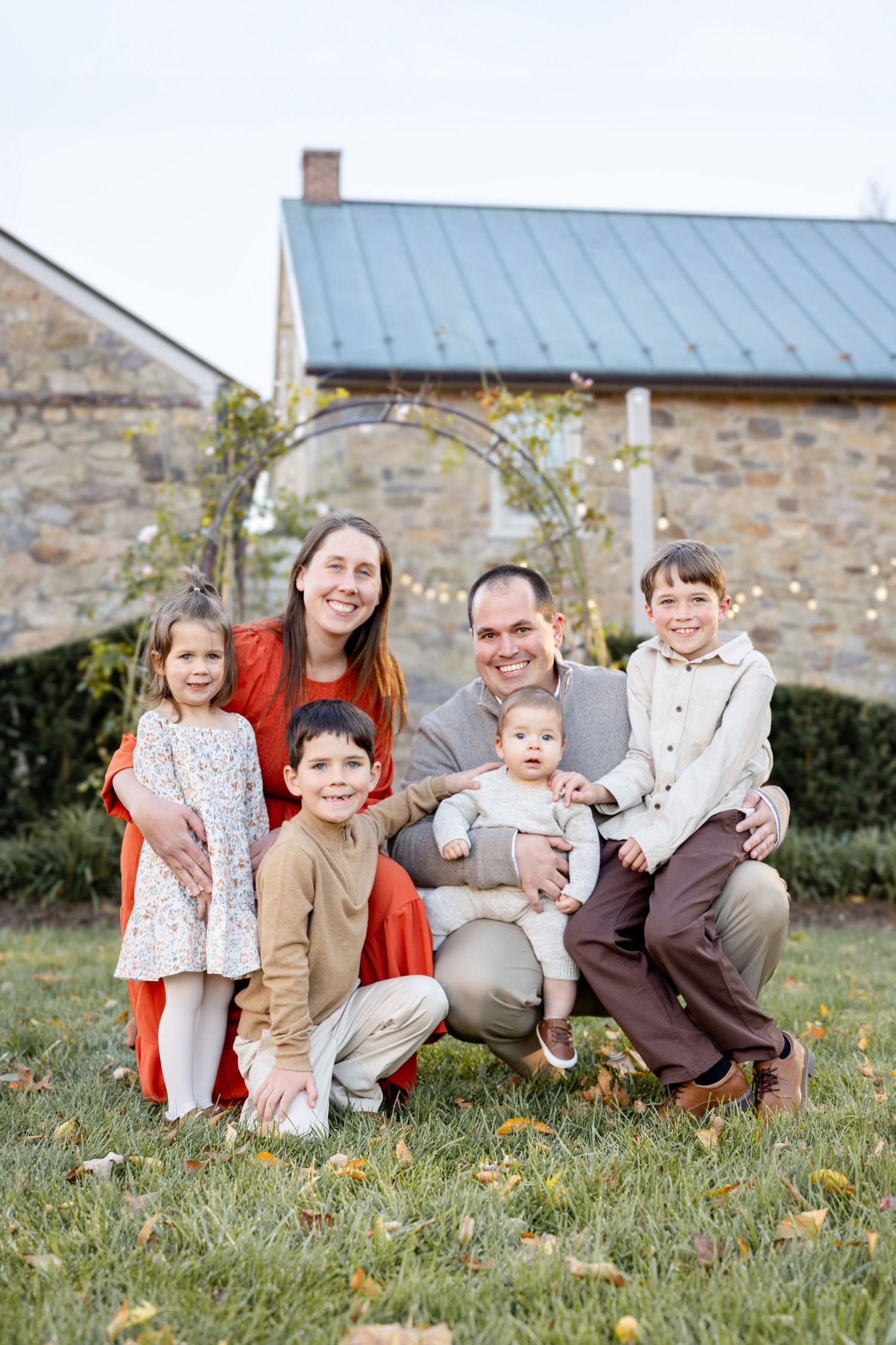 A smiling family of six poses outdoors on a lawn in front of a stone building, dressed in coordinated fall outfits.