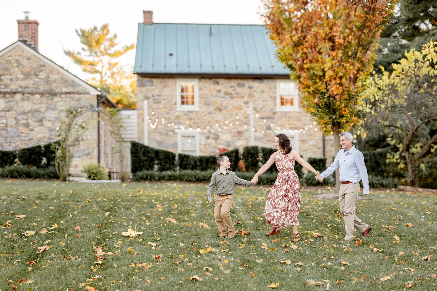 Family holding hands and walking on a green lawn with autumn trees and a historic stone house in the background.