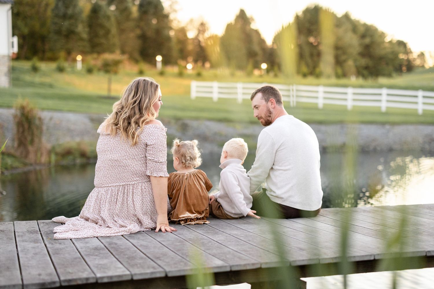 A family sits together on a wooden dock by a pond at sunset with white fences and trees in the background.