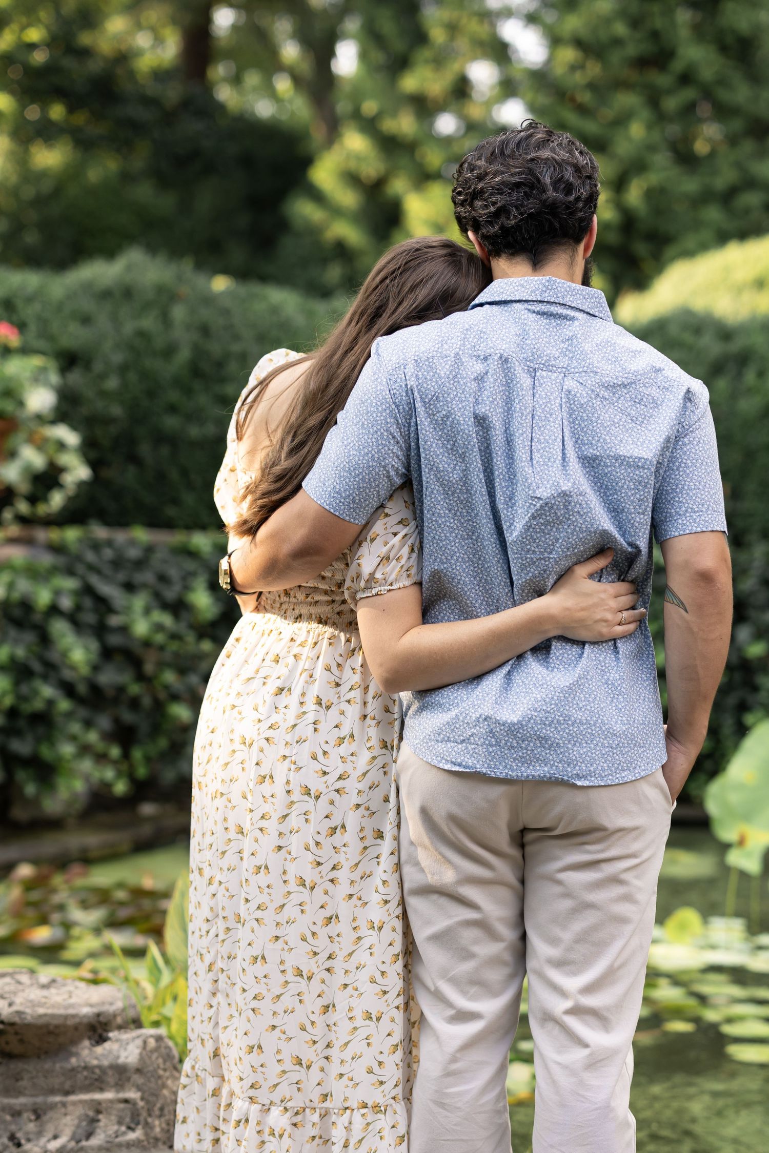 A couple embraces in a lush garden, viewed from behind, woman in floral dress leaning on man in blue shirt.