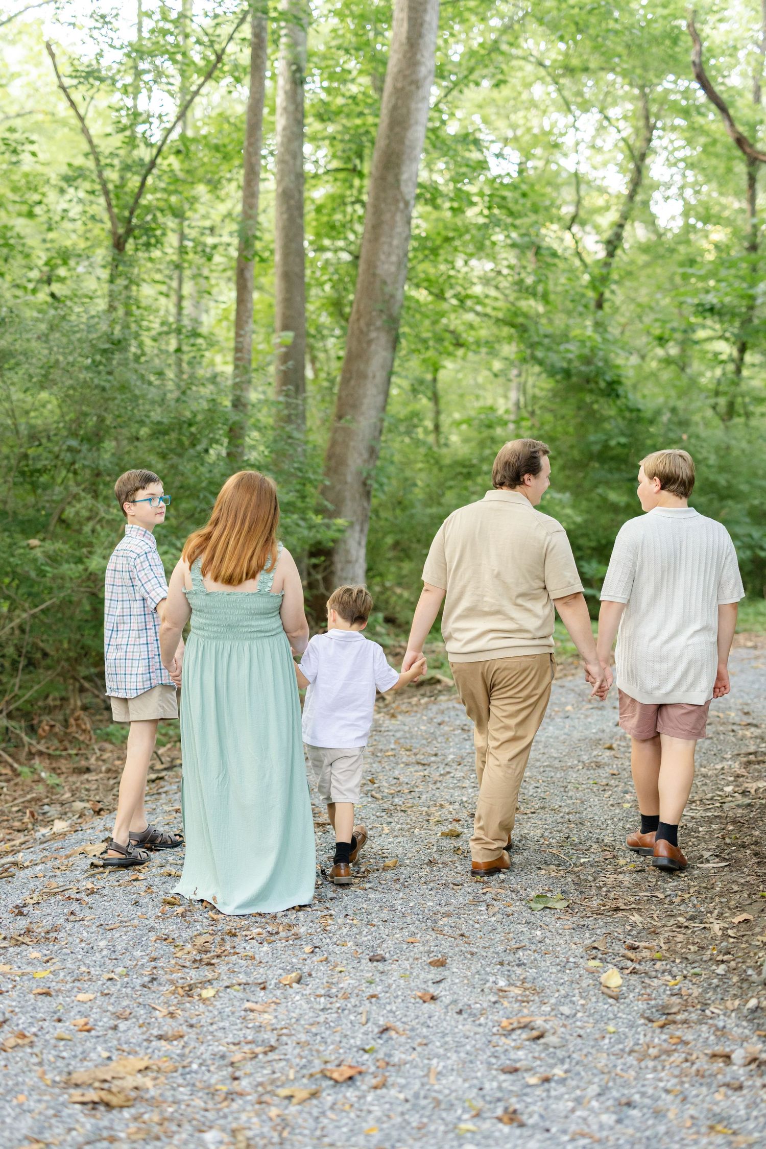 Family of five walking hand-in-hand on a gravel path through lush green woods, viewed from behind.