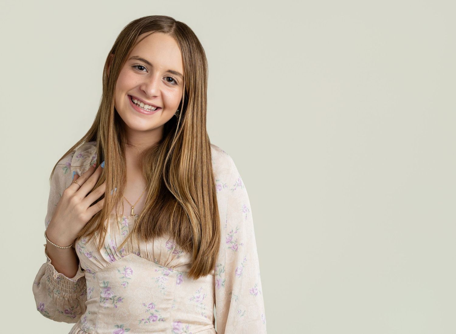 Smiling teenage girl with long brown hair and braces wearing a floral blouse against a light green background.