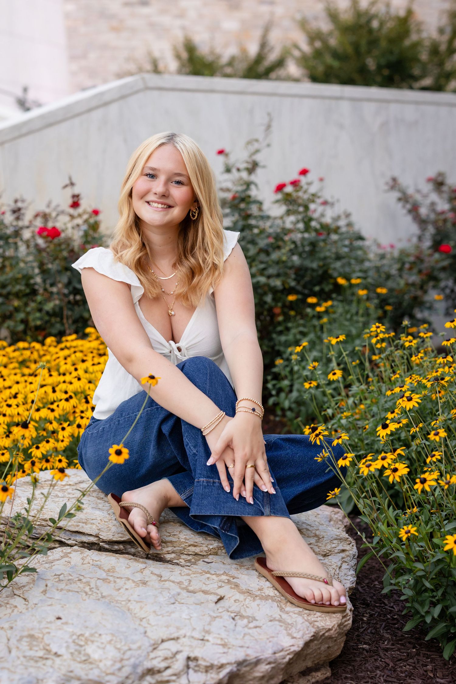A high school senior girl in blue jeans sits on a rock surrounded by vibrant yellow Black-eyed Susan flowers and red blooms in a garden.