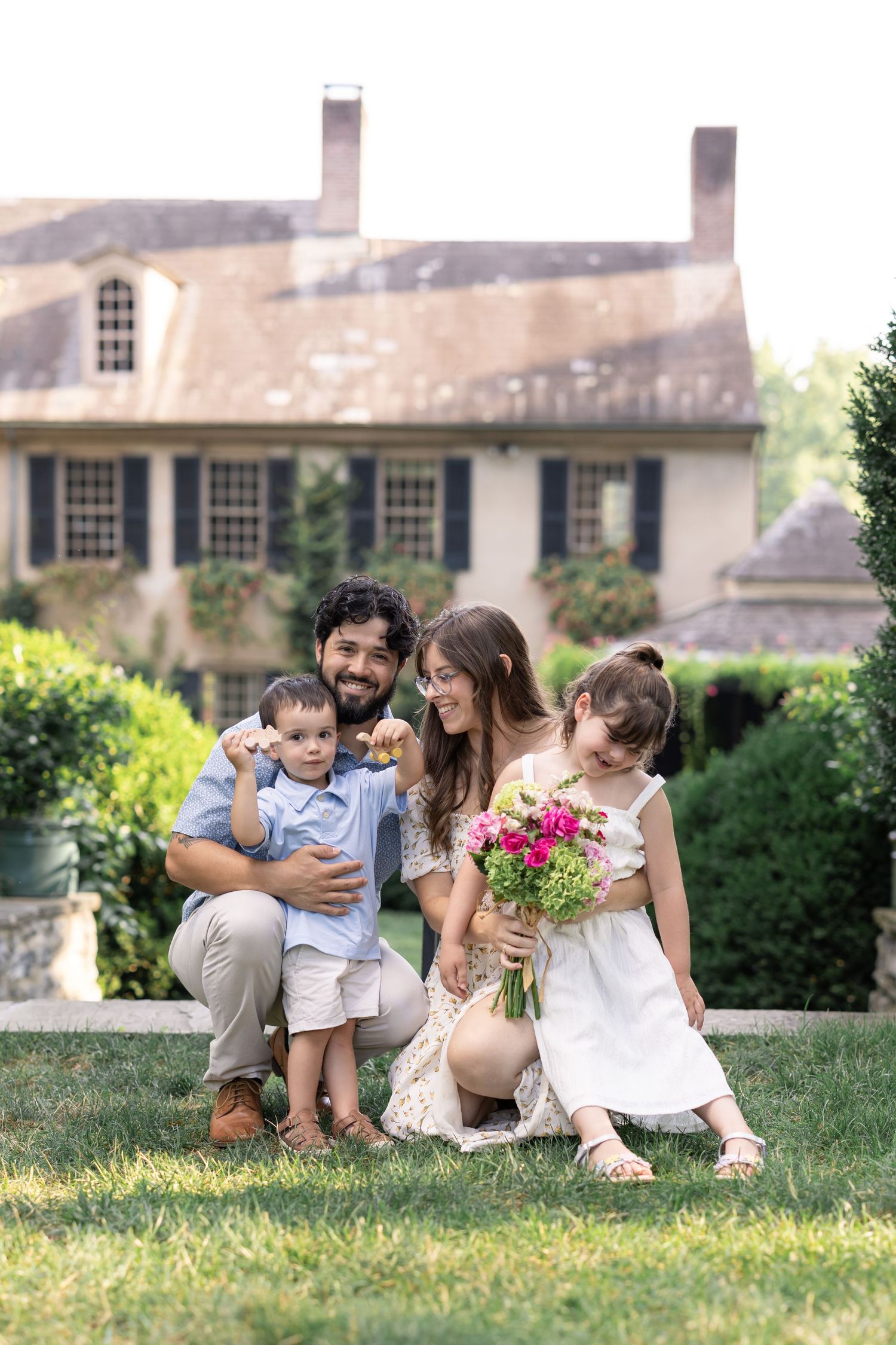 Happy family of four posing on green lawn in front of a historic estate, mother holding pink flower bouquet.