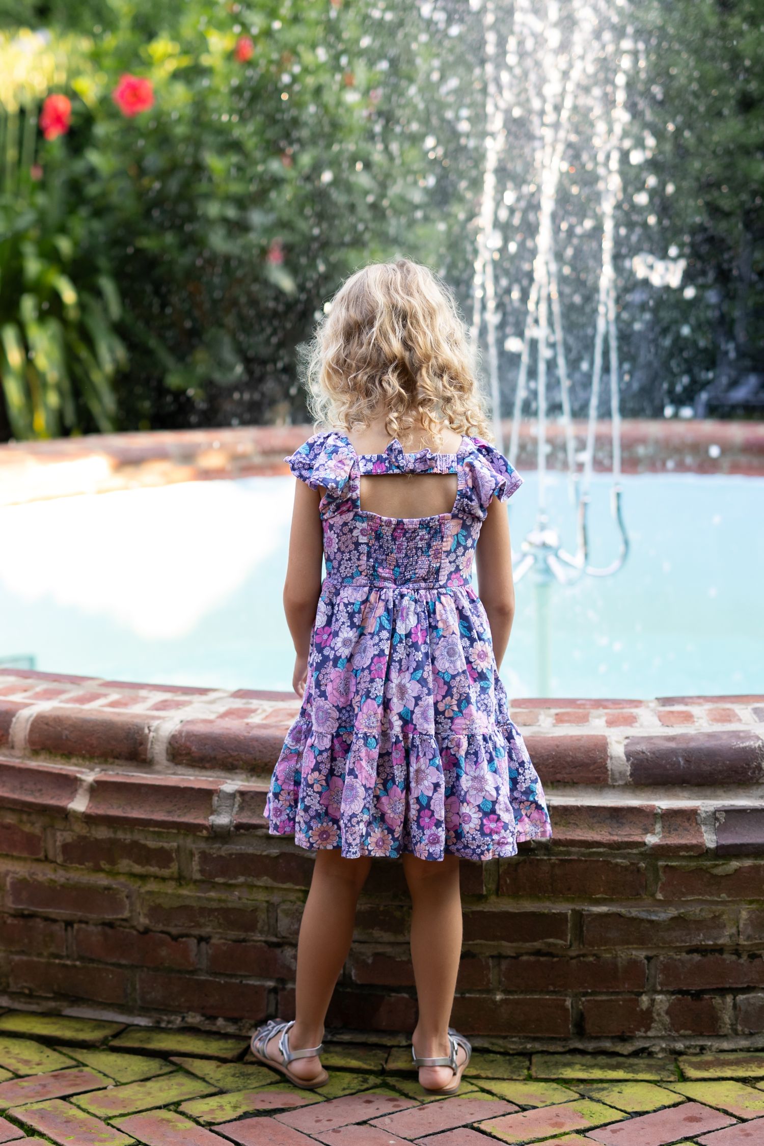Young child in a purple floral dress stands at edge of fountain with water jets spraying in garden setting.