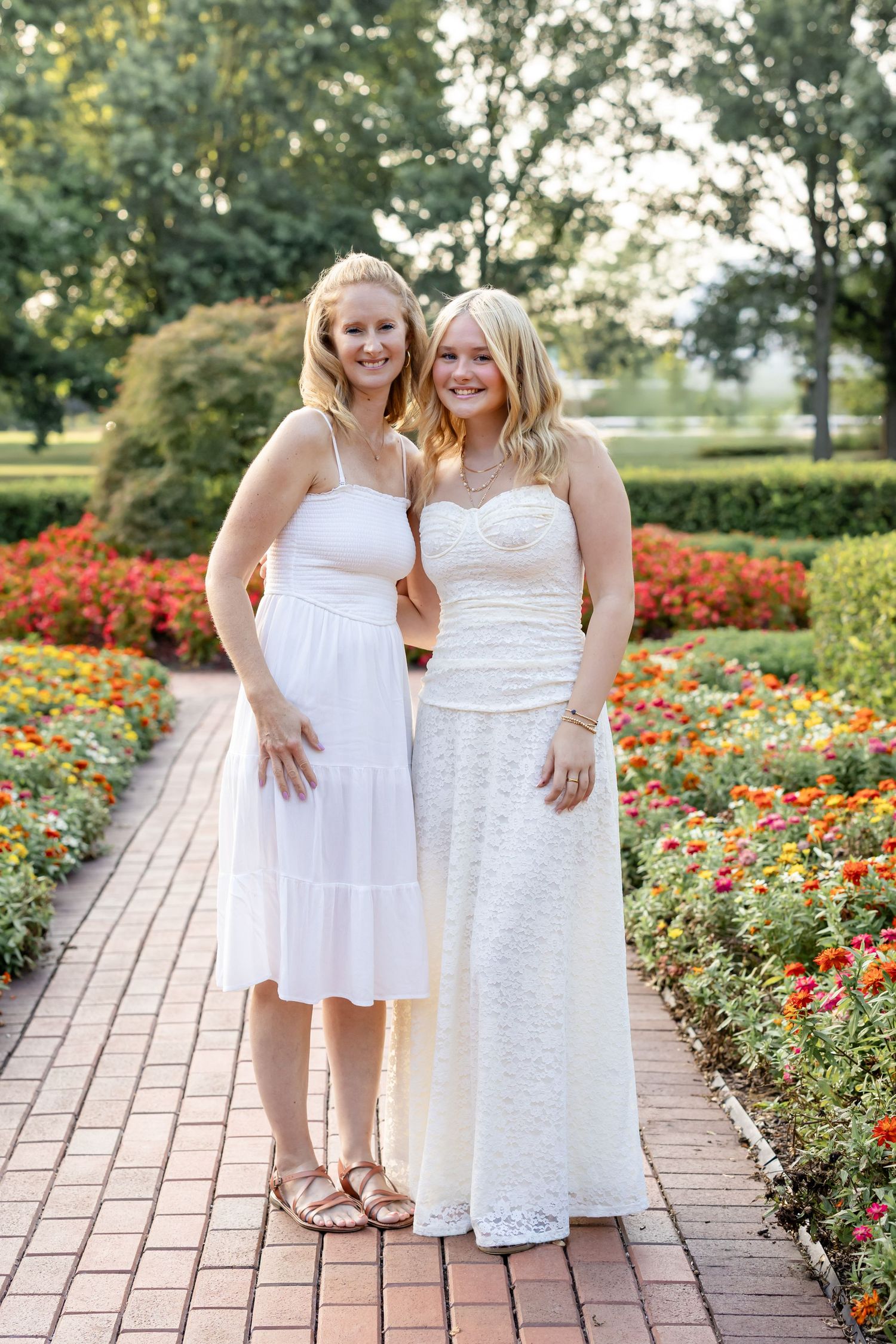 A high school graduate with her mom in white dresses posing together on a brick path surrounded by colorful flower gardens.