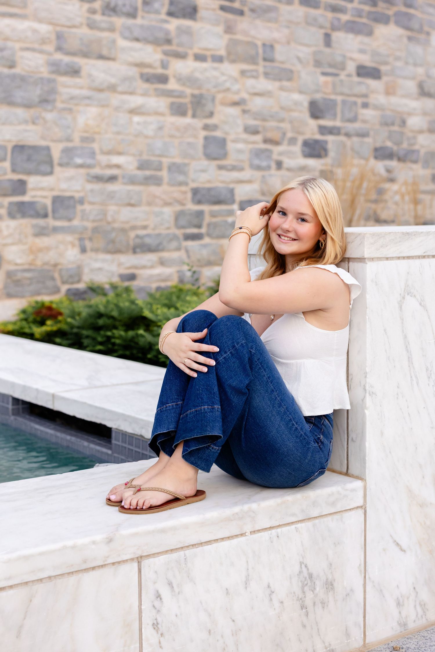 A high school senior girl in blue jeans and white top lounges on a modern stone bench against a brick wall backdrop.