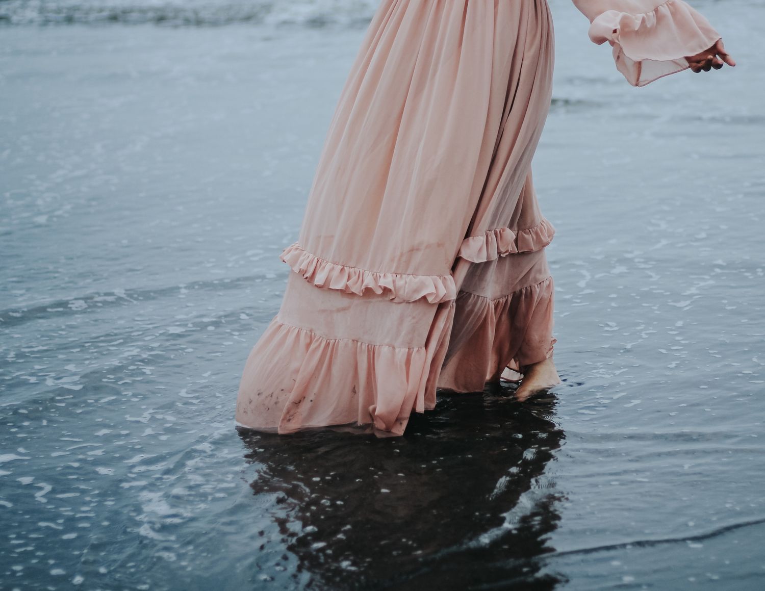 A high school senior girl wearing a flowing pink dress stands in shallow water creating a dreamy reflection at sunset.