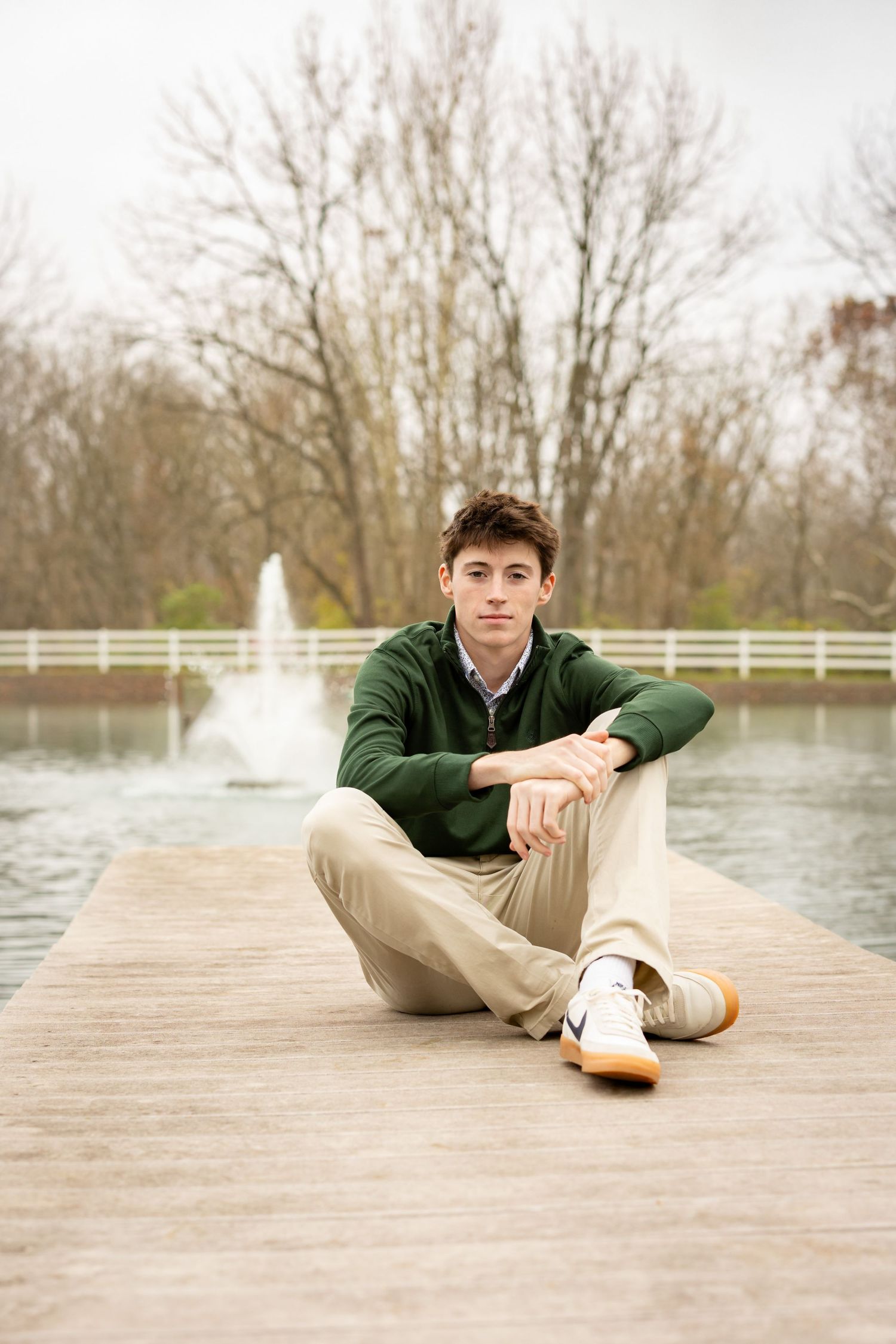 A high school boy in green sweater and khaki pants lying on wooden dock by water on cloudy day.