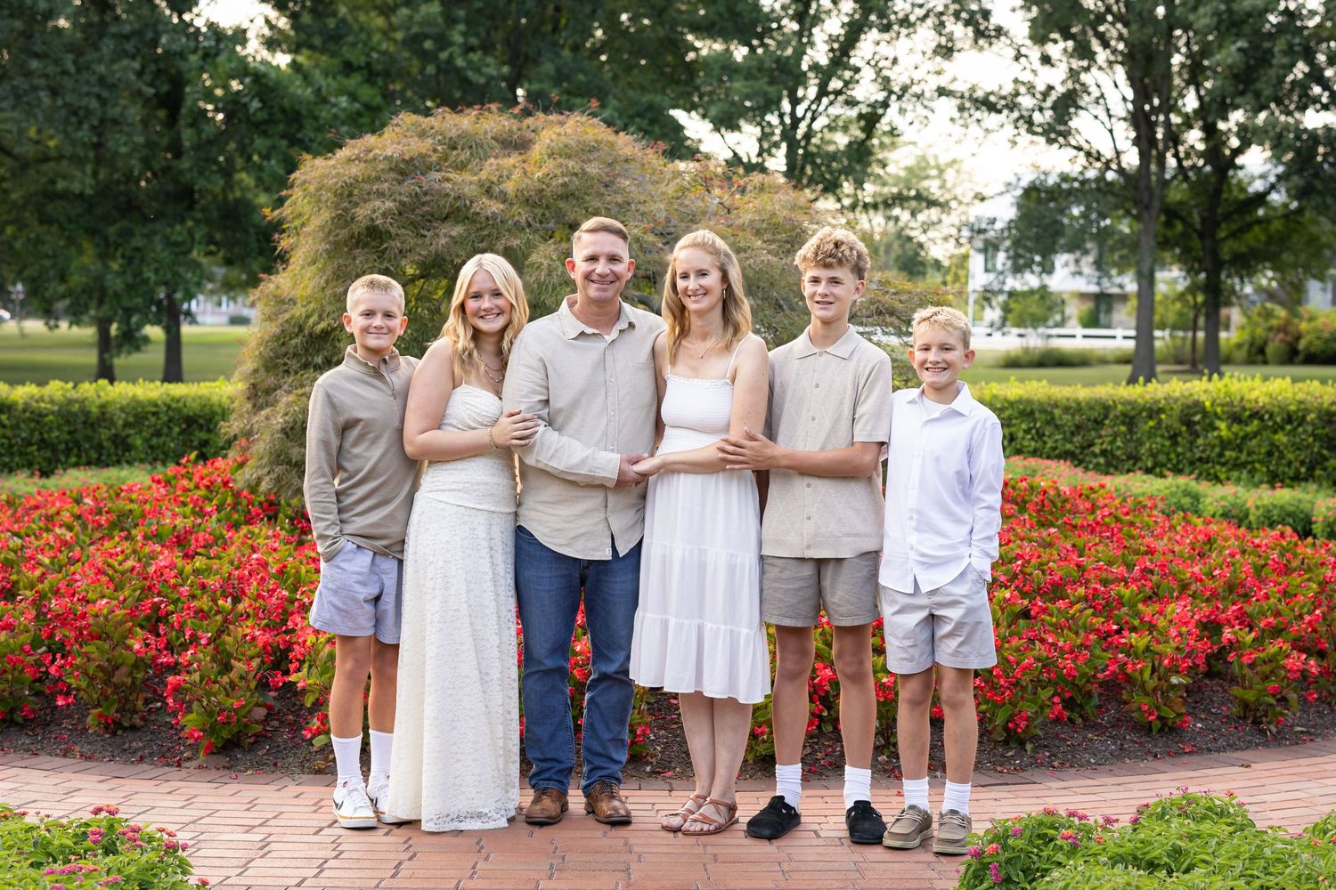 Family of six posing together in a garden with red flowers and green trees in the background.