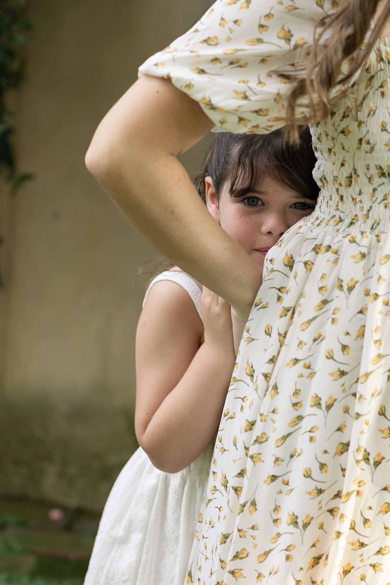Young girl in white dress peeking shyly from behind mother's floral dress outdoors.