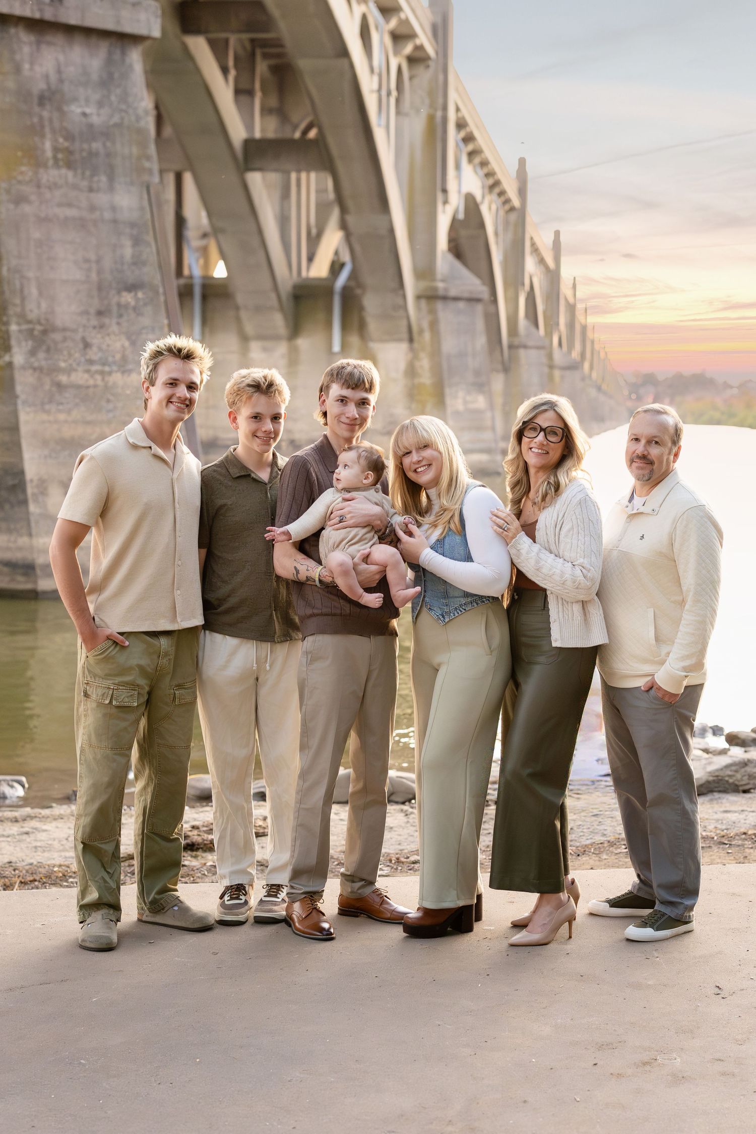 Multi-generational family of seven poses under a bridge at sunset, including a woman holding a baby.