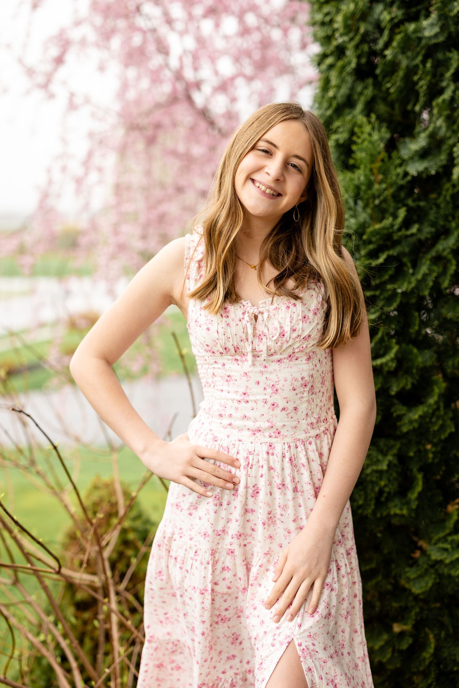 Young smiling teen girl in pink floral sundress posing outdoors near blooming cherry blossom tree in spring garden.