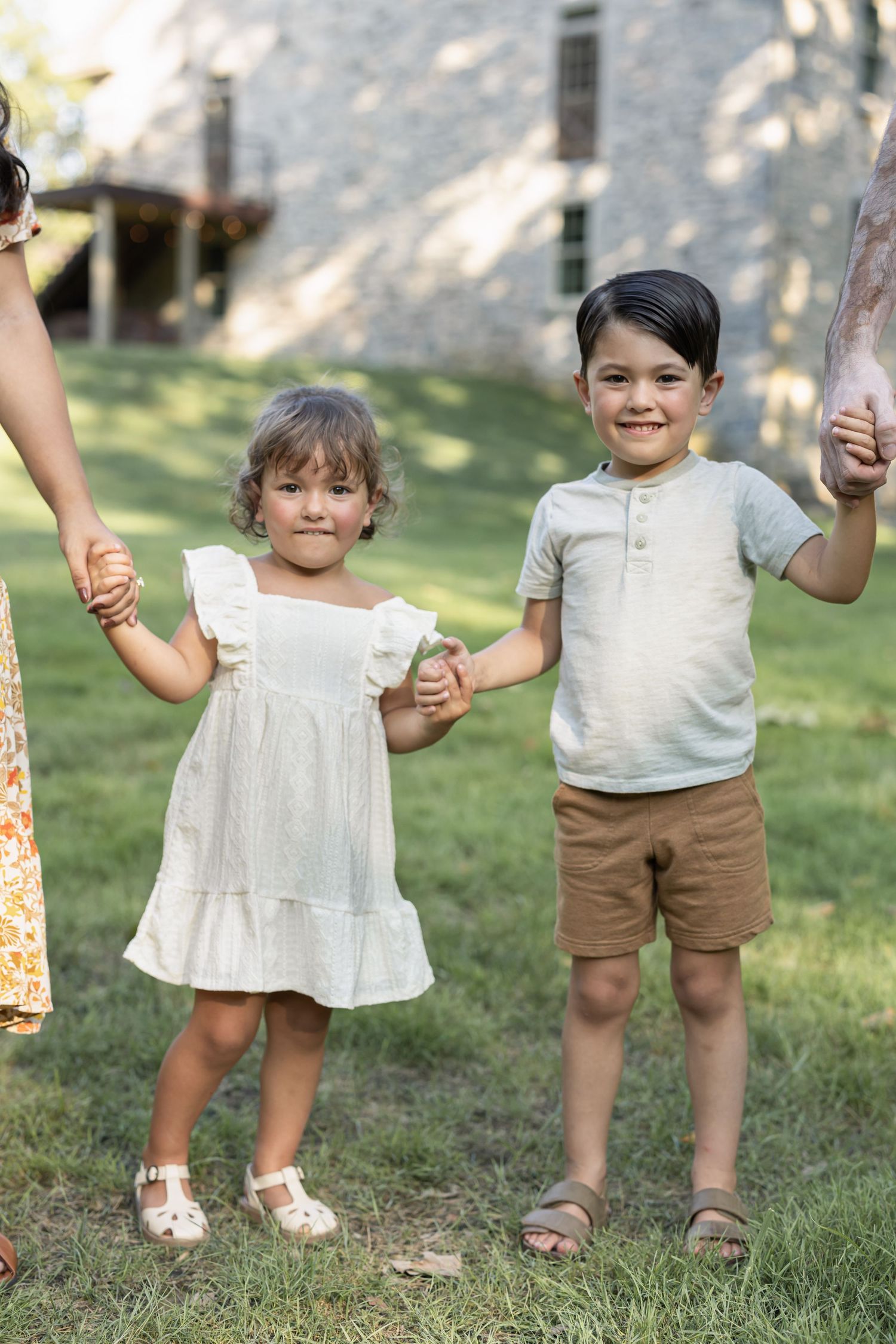 Two young children holding hands with adults outdoors on green grass near a stone building on a sunny day.