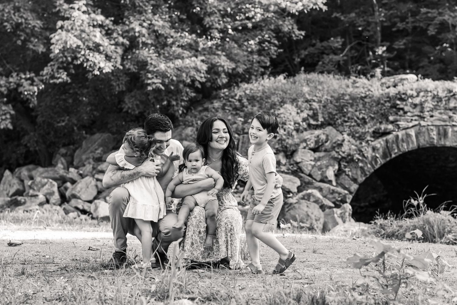 Black and white family photo outdoors near stone bridge, parents and children laughing together in a park setting.