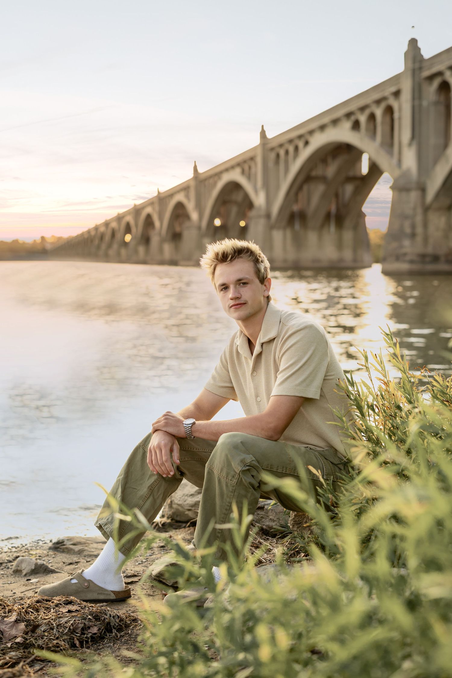 High school senior portrait of a boy sitting by a stone bridge at sunset, surrounded by riverside vegetation and warm golden light.