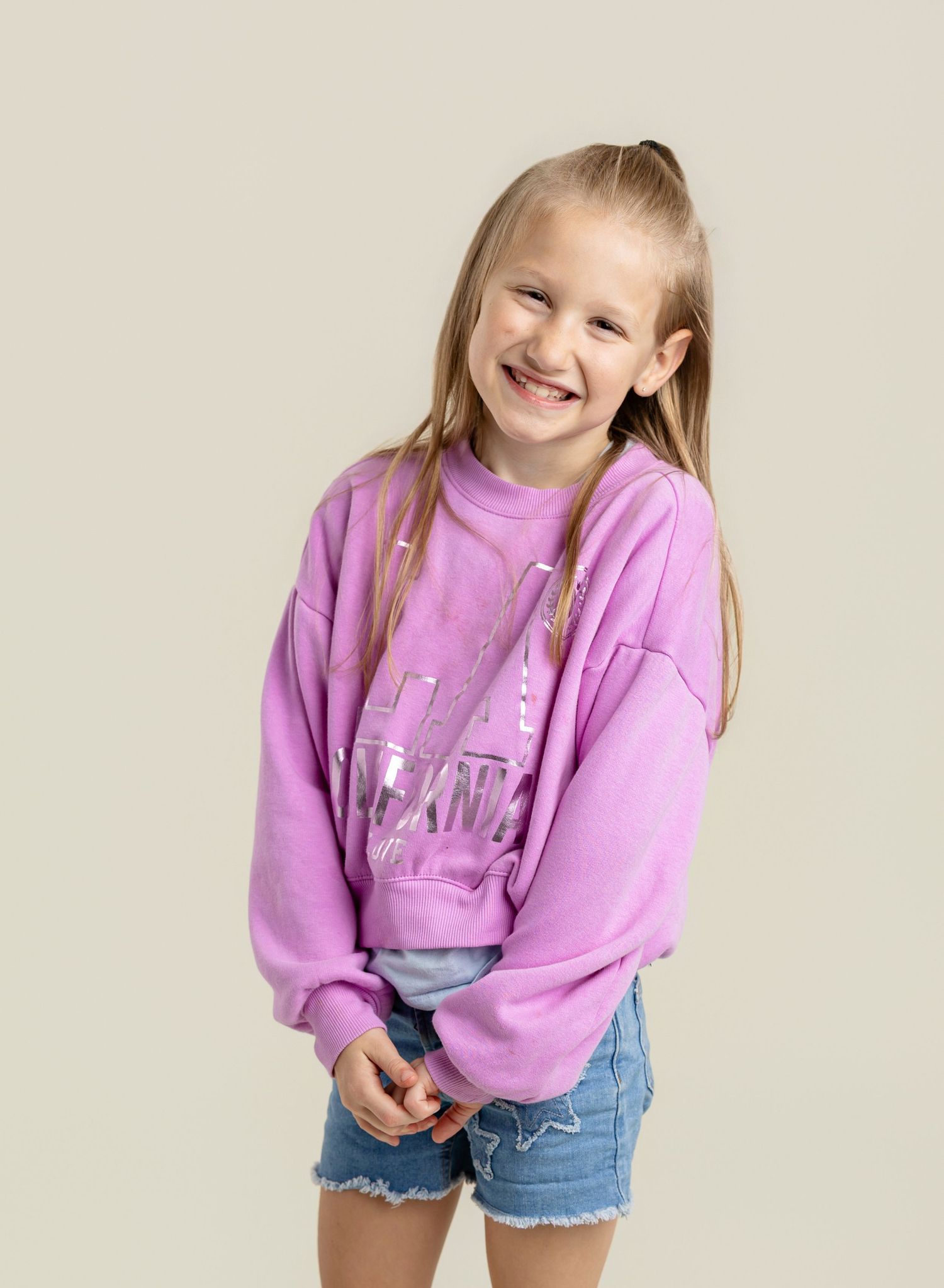 Smiling girl wearing a purple oversized sweatshirt and denim shorts, posing cheerfully against a neutral background.